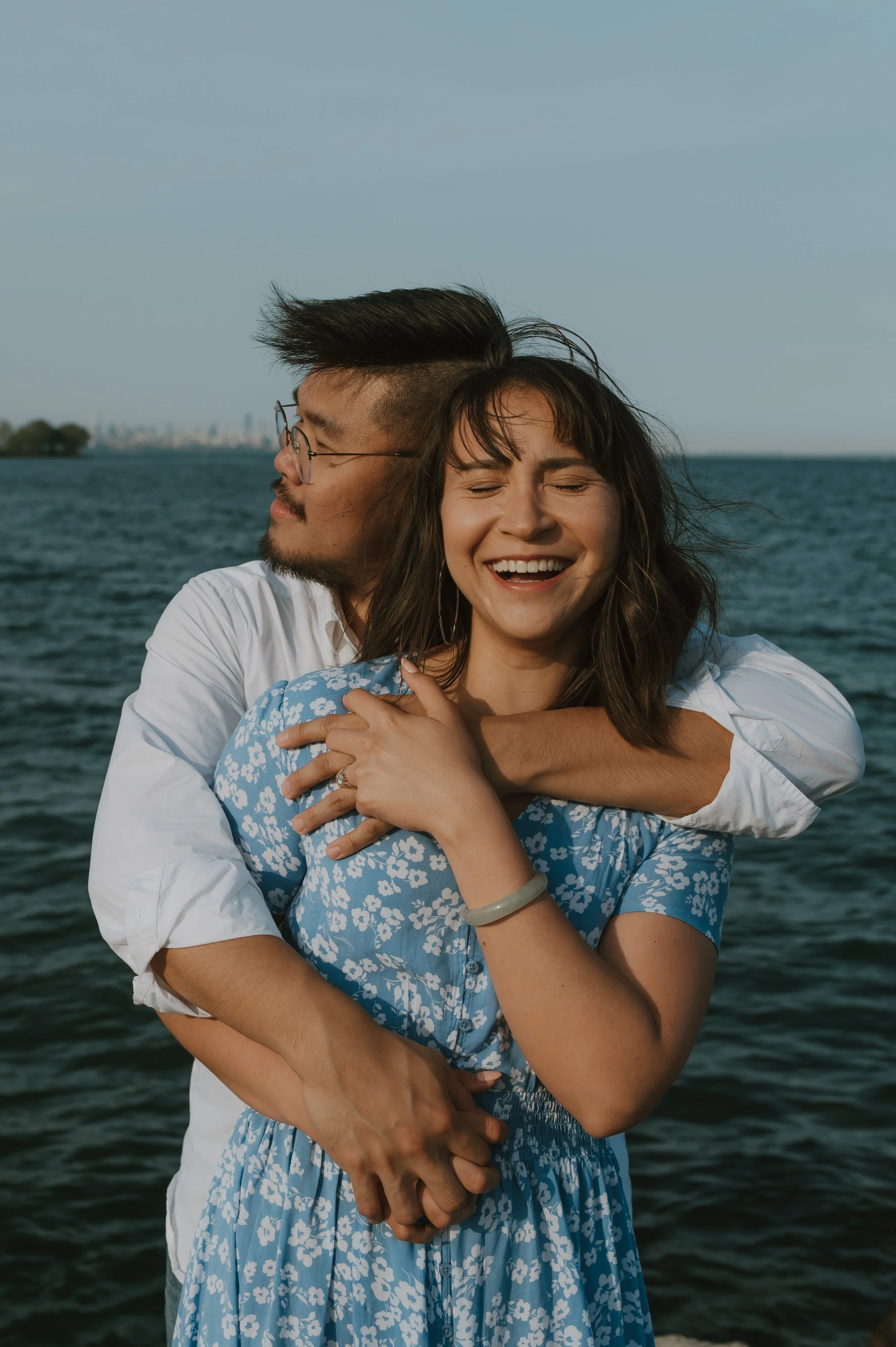A couple hugging by the water, the woman smiling with eyes closed and the man kissing her on the cheek, with city skyline in the background.
