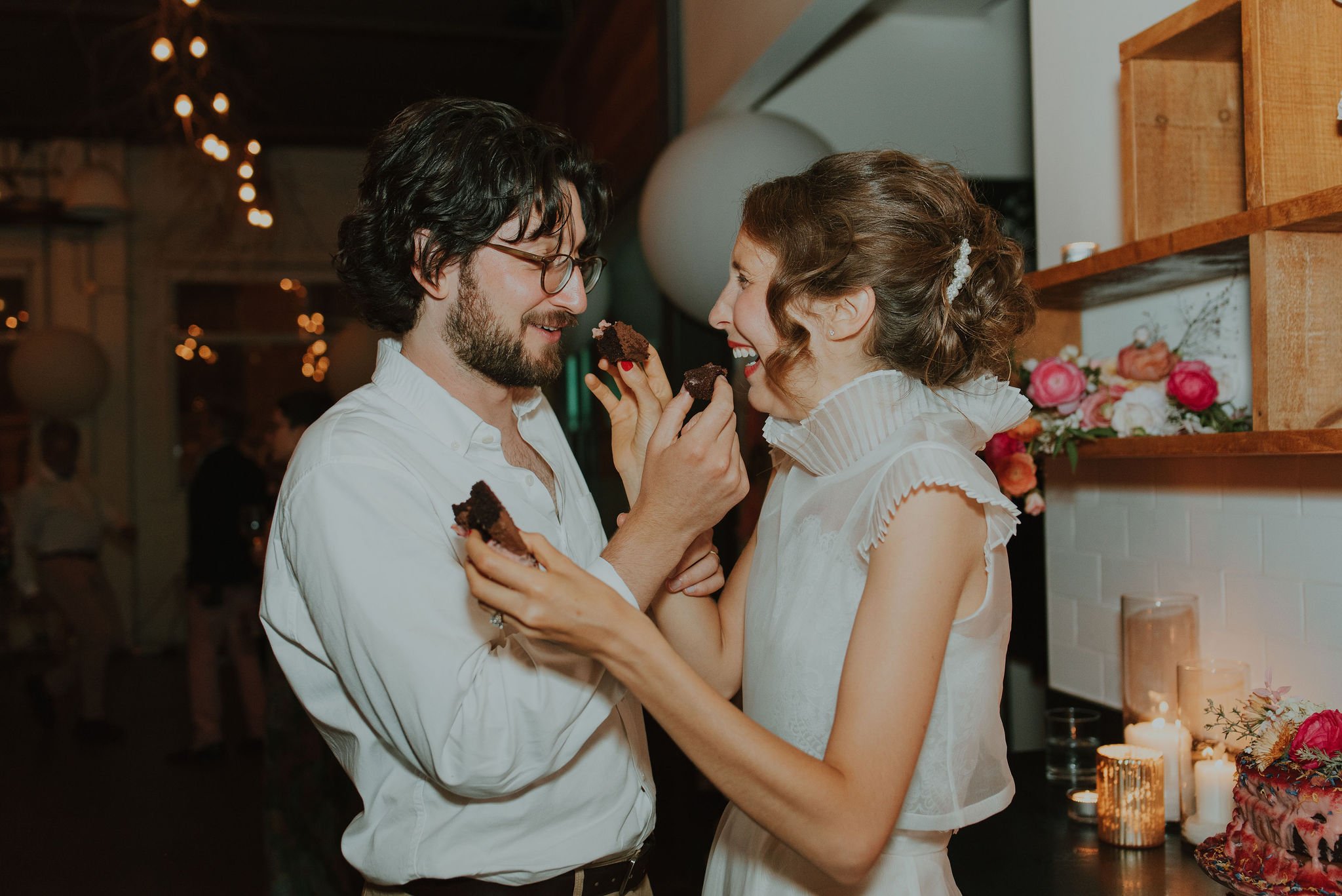 A couple smiling and sharing chocolate cake at a celebration, surrounded by candles and flowers.
