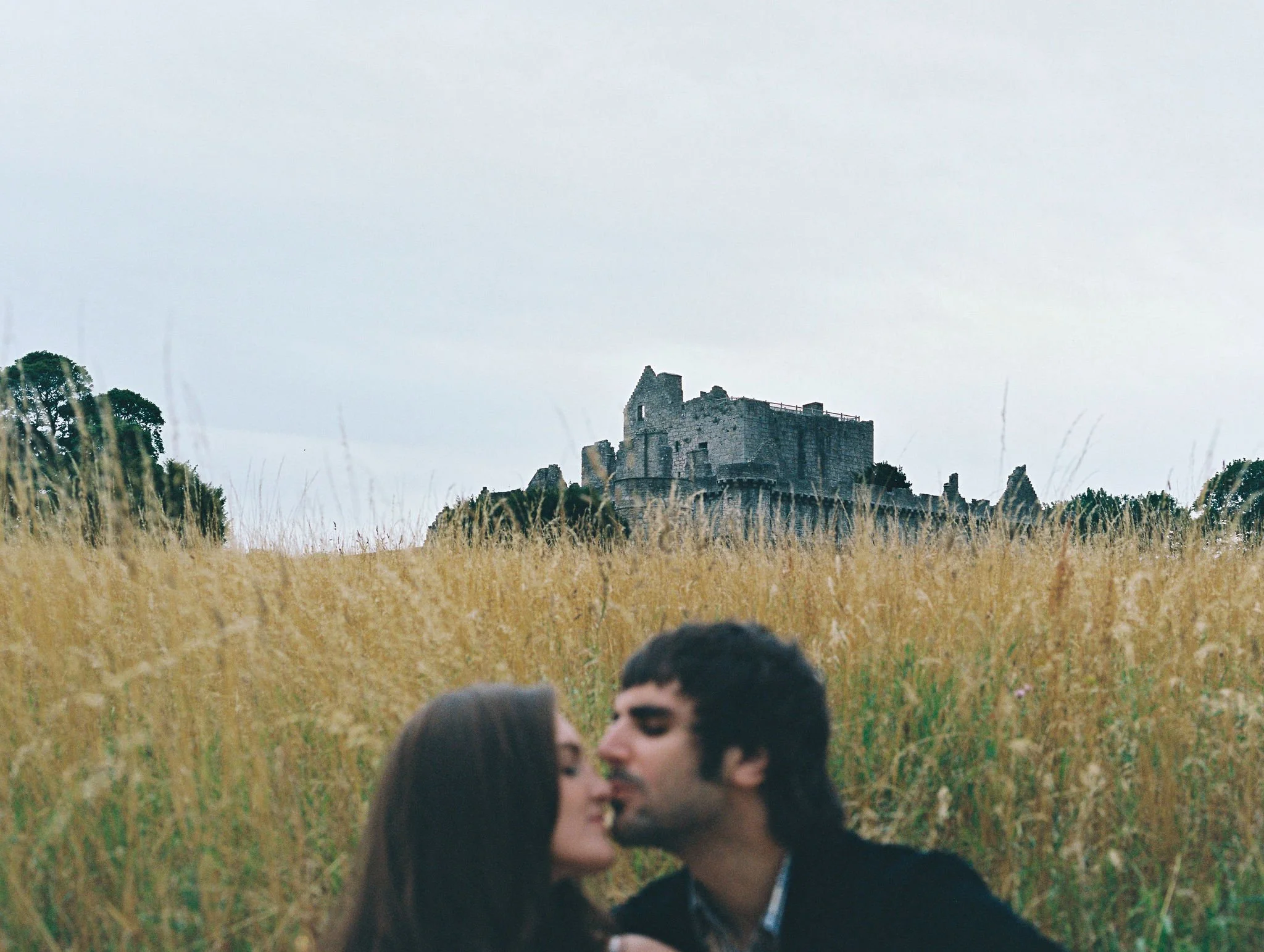 A couple kissing in a field of tall, golden grass with an ancient stone castle in the background under an overcast sky.