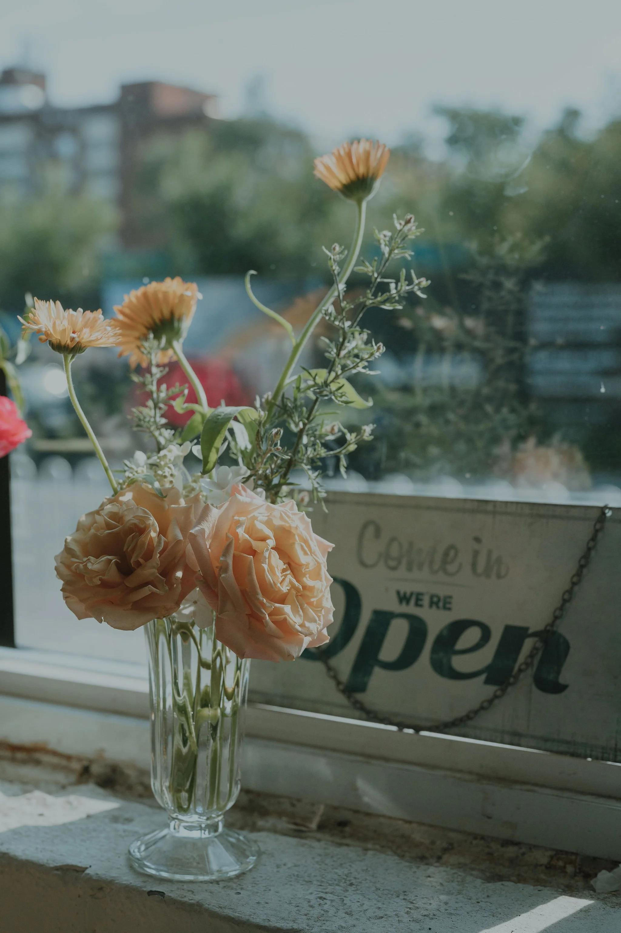 A glass vase with peach-colored roses and yellow daisies on a windowsill, with a sign in the background that says 'Come in we're open'.