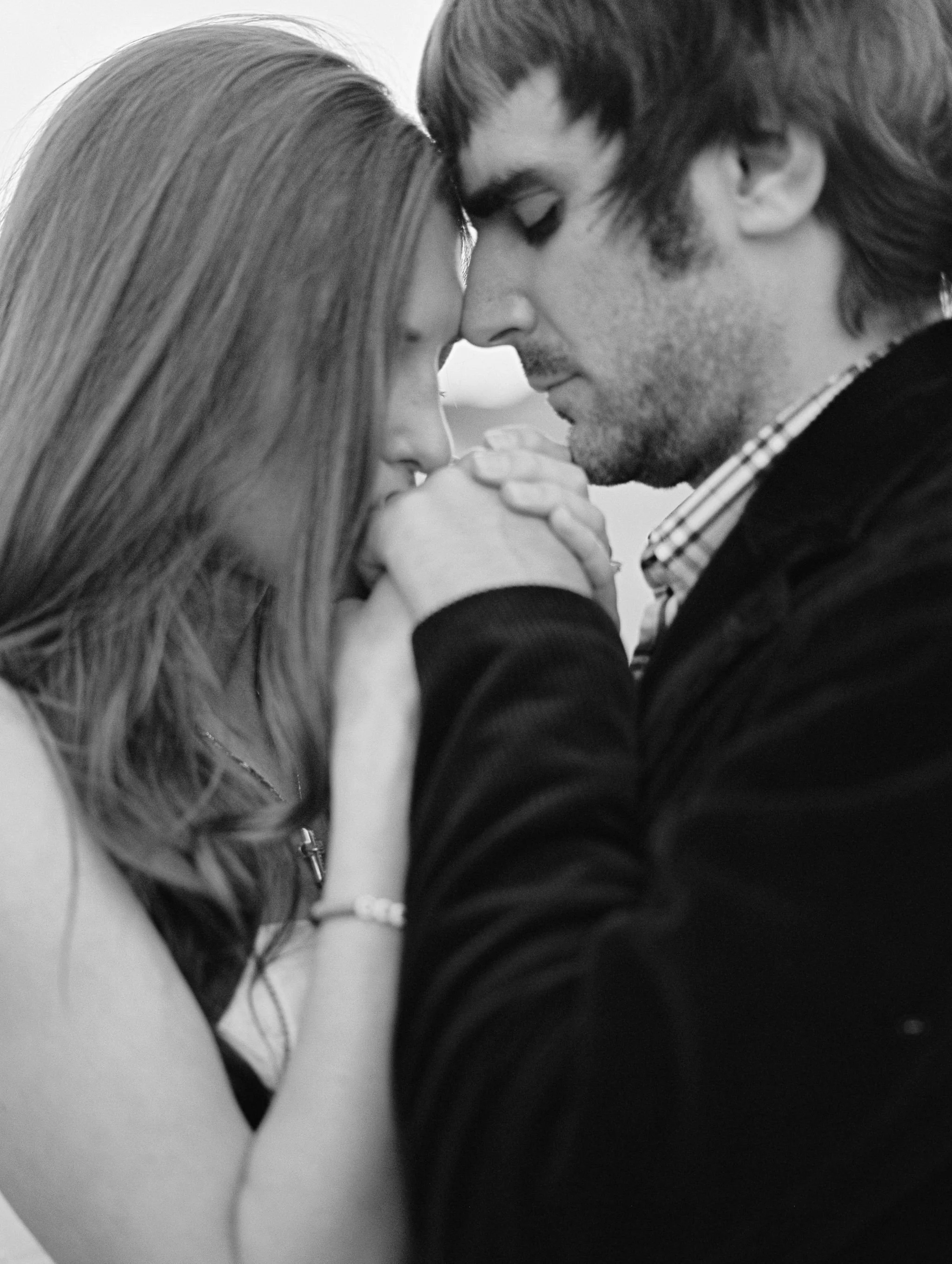 A black and white close-up photo of a man and woman with eyes closed, forehead to forehead, holding hands and touching foreheads, sharing an intimate moment.