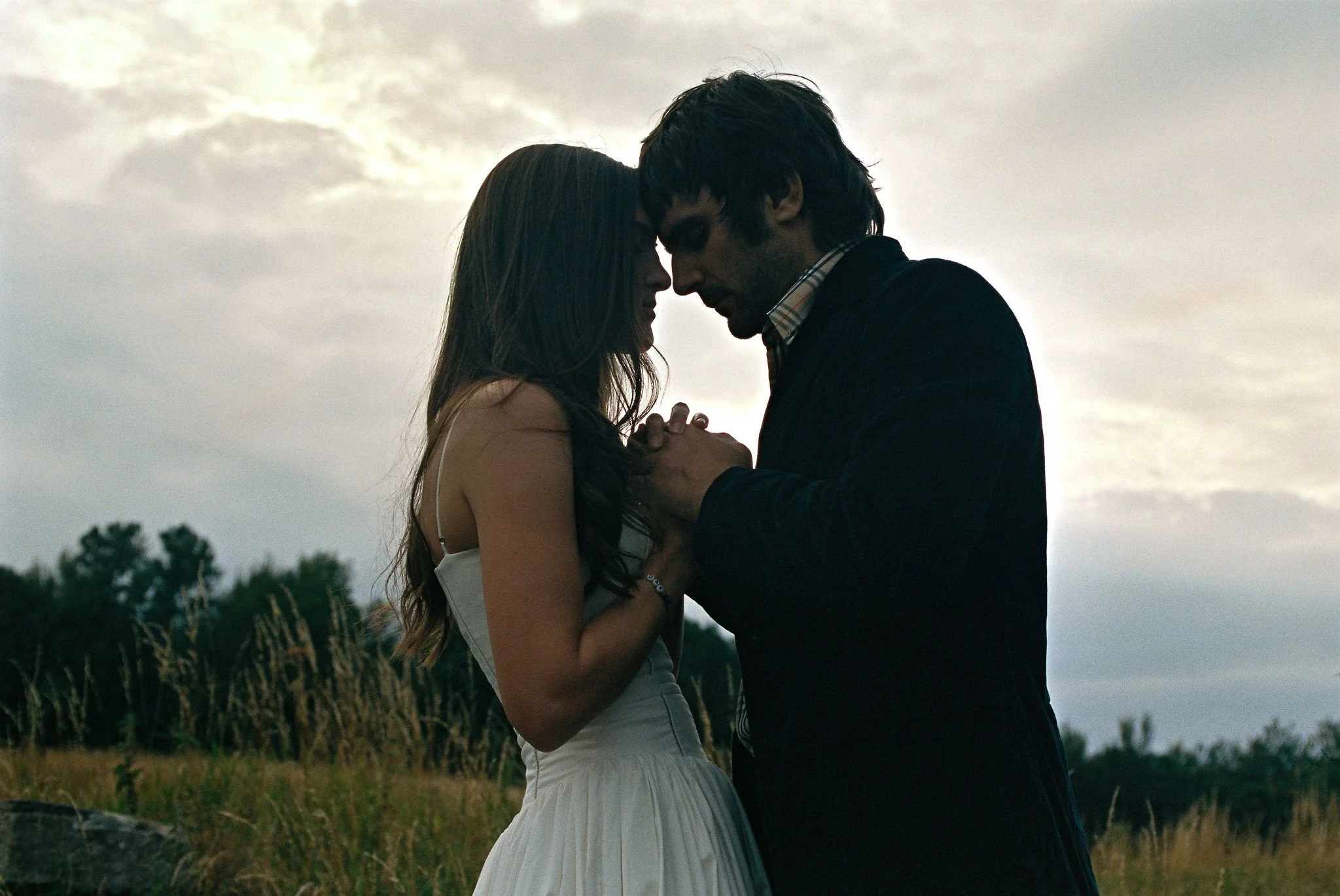 A man and woman are standing outdoors with their foreheads touching, holding hands, in a tender moment at sunset or dusk.