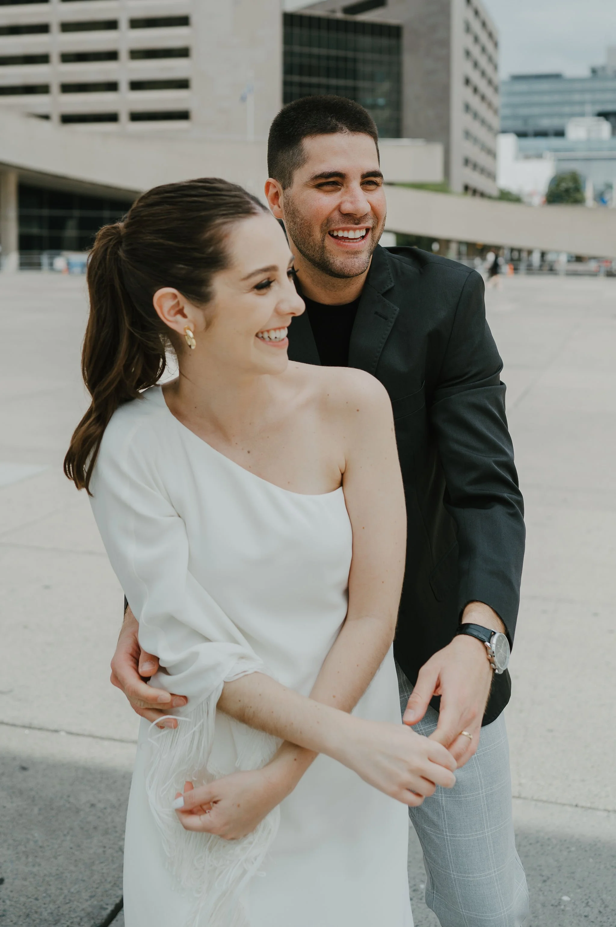 A smiling couple standing outdoors in an urban setting, with the man gently embracing the woman from behind.