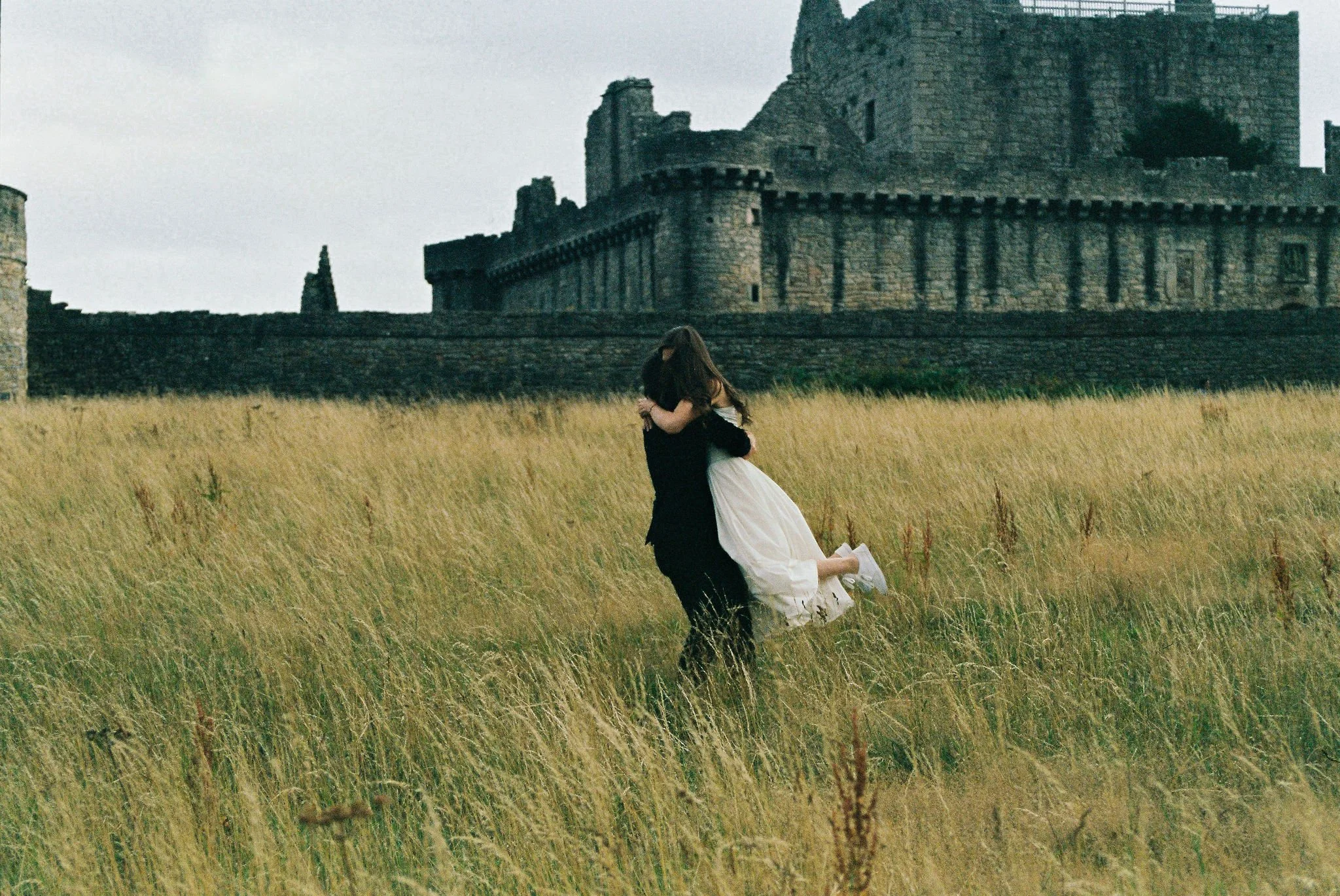 A couple embracing in a grassy field with an ancient stone castle in the background.