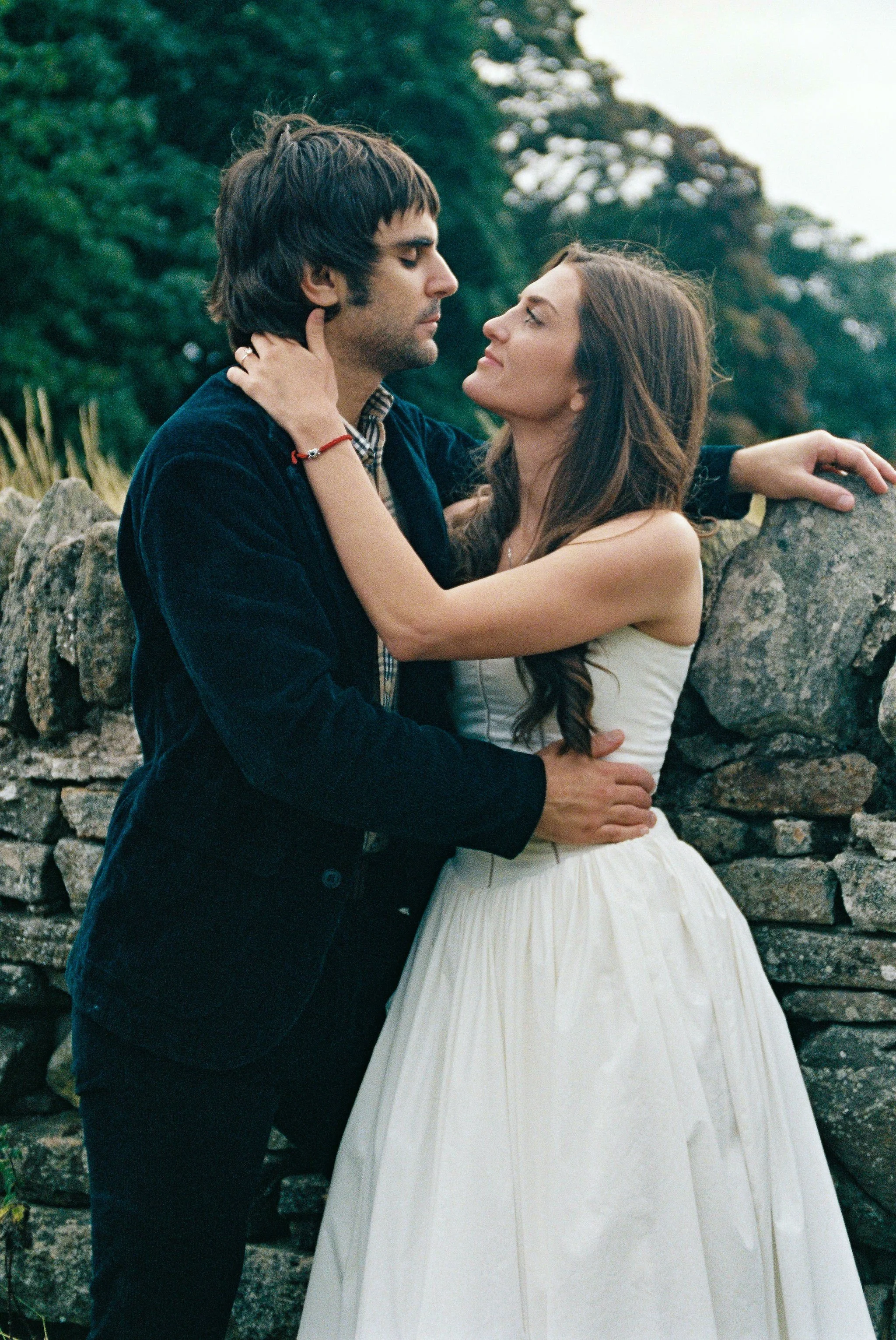 A man and woman embrace outdoors near a stone wall, looking into each other's eyes with trees in the background.