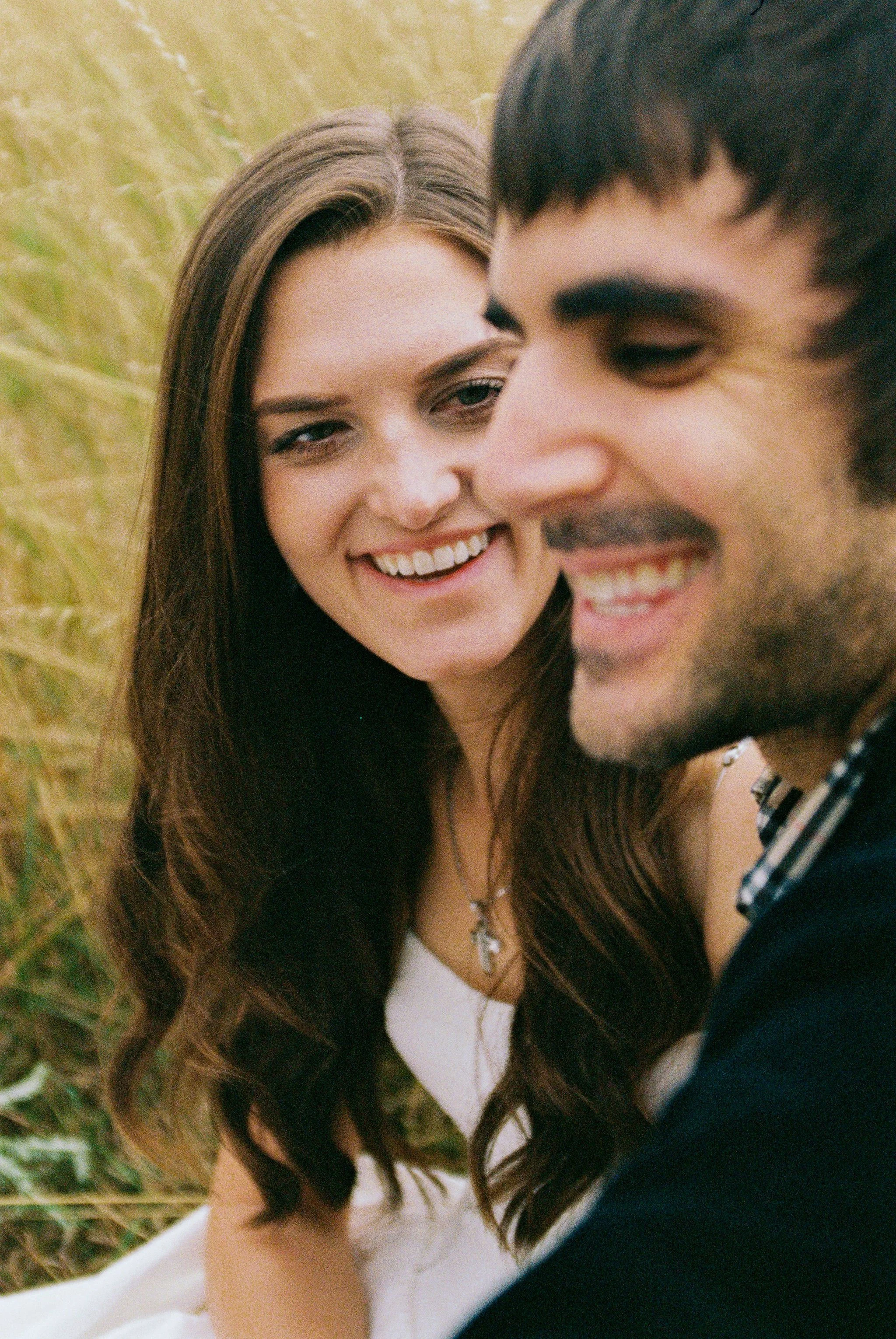 A smiling couple outdoors in a field with tall grass, close-up of their faces showing happiness.