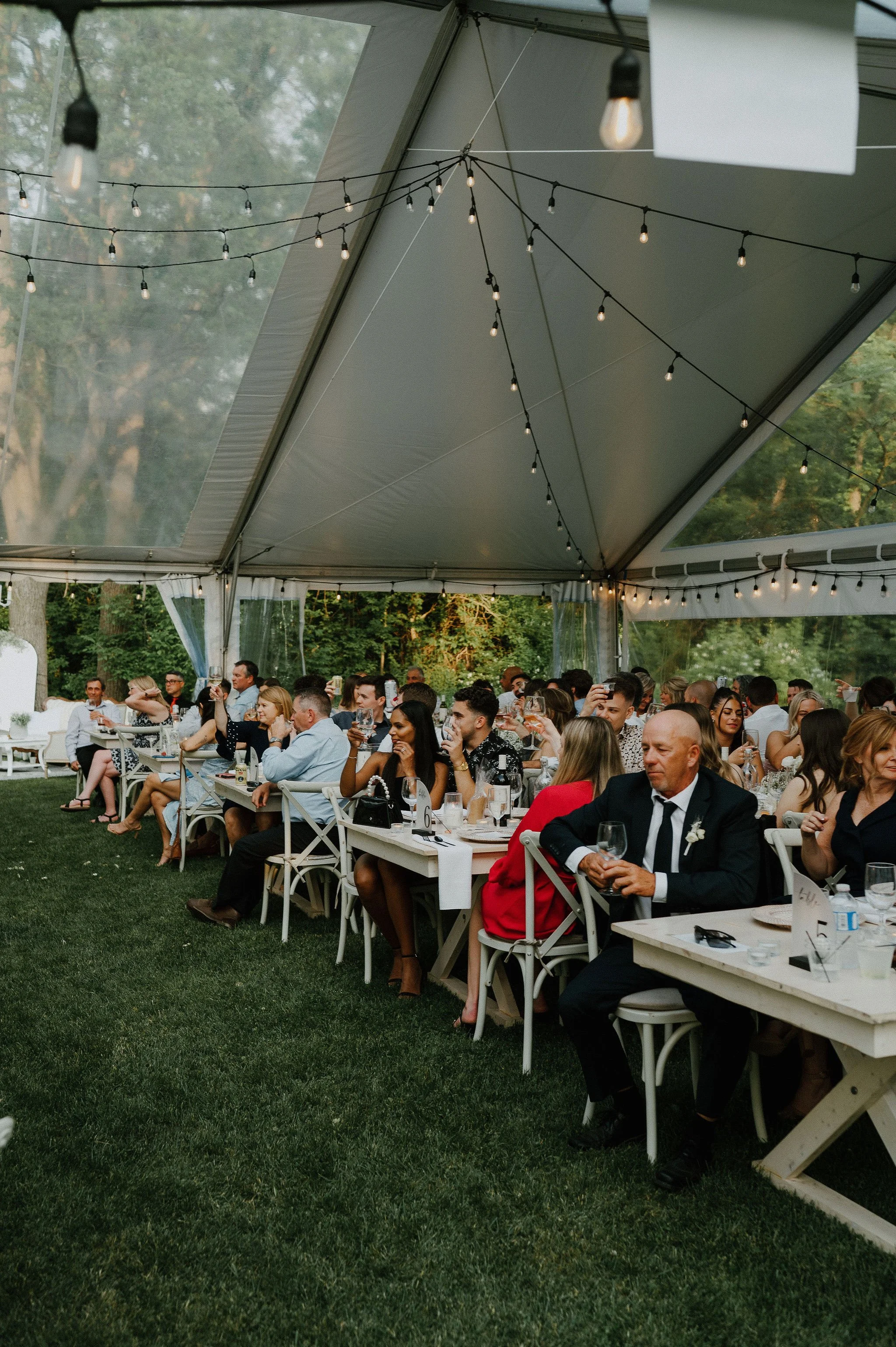 People sitting at tables under a large outdoor tent with string lights, attending a celebratory gathering or wedding reception.
