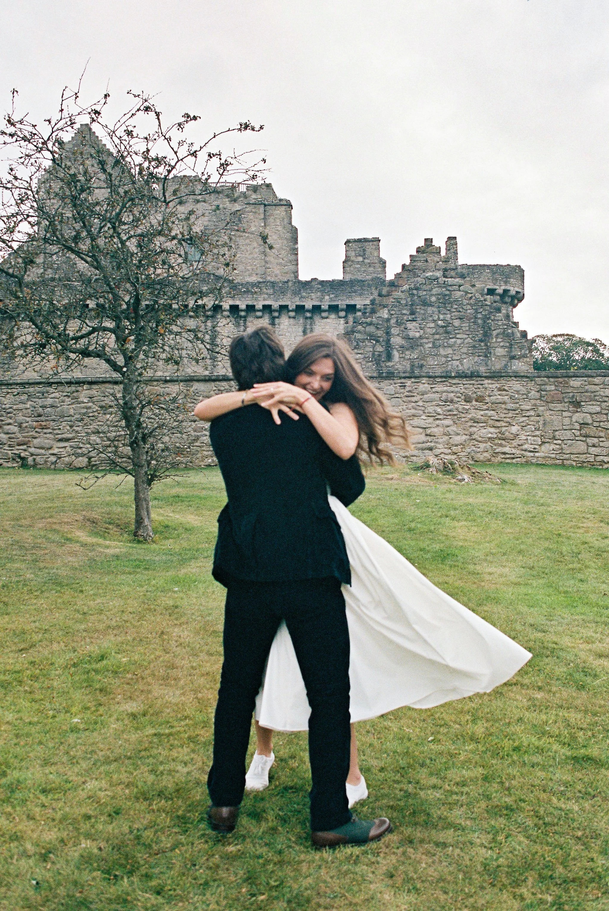 A couple dancing outdoors on a grassy area, with a woman in a white dress and a man in dark clothing, embracing in front of a stone castle and a leafless tree on a cloudy day.