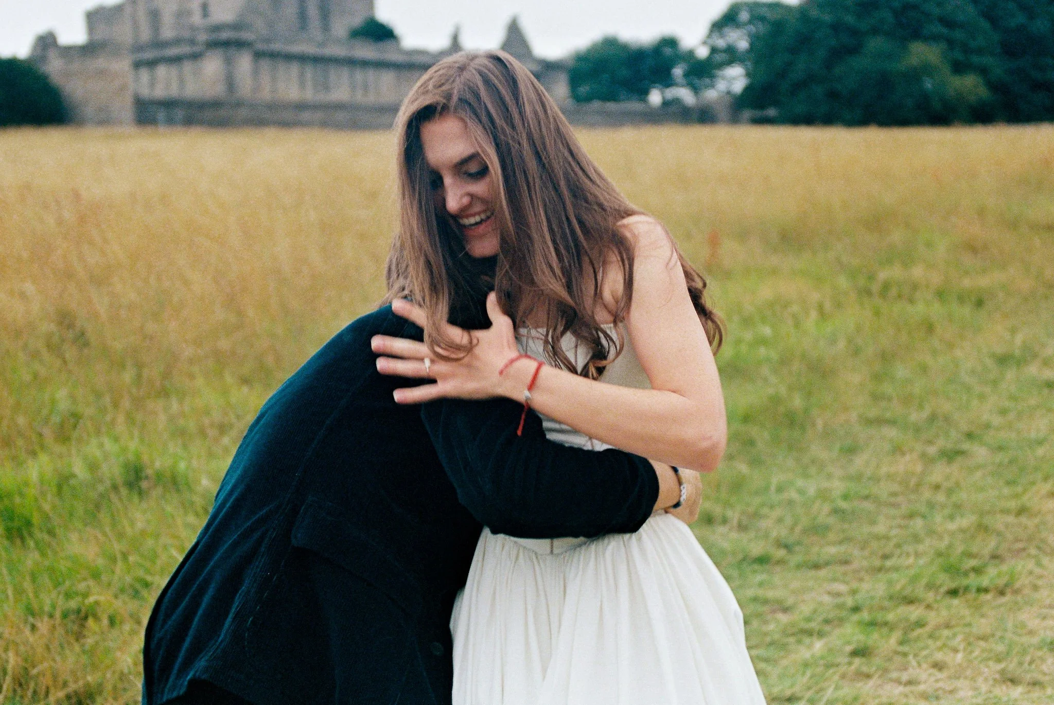 A woman with long brown hair wearing a white dress hugging a person in a dark jacket in a grassy field with a castle in the background.