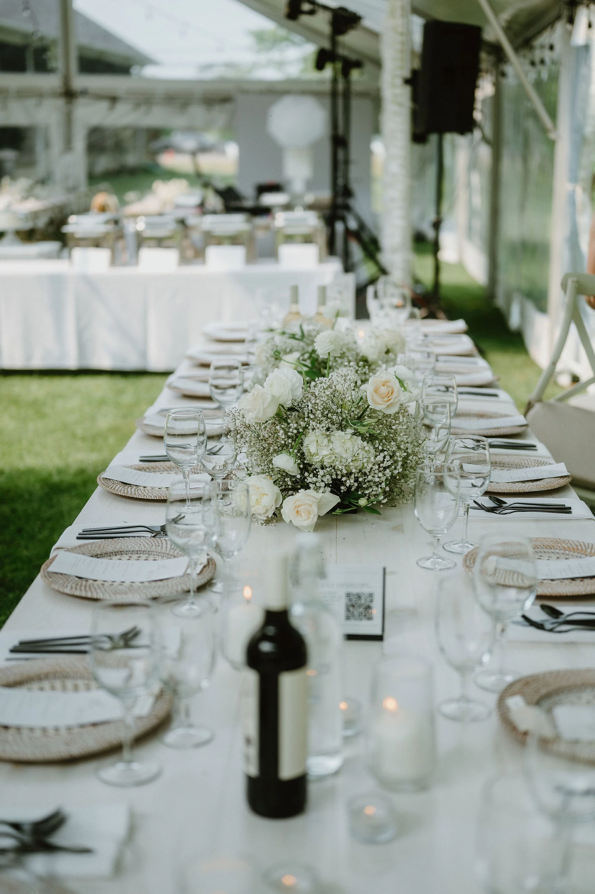 A elegantly set outdoor dining table decorated with white flowers, wine glasses, and place settings, under a tent or canopy.