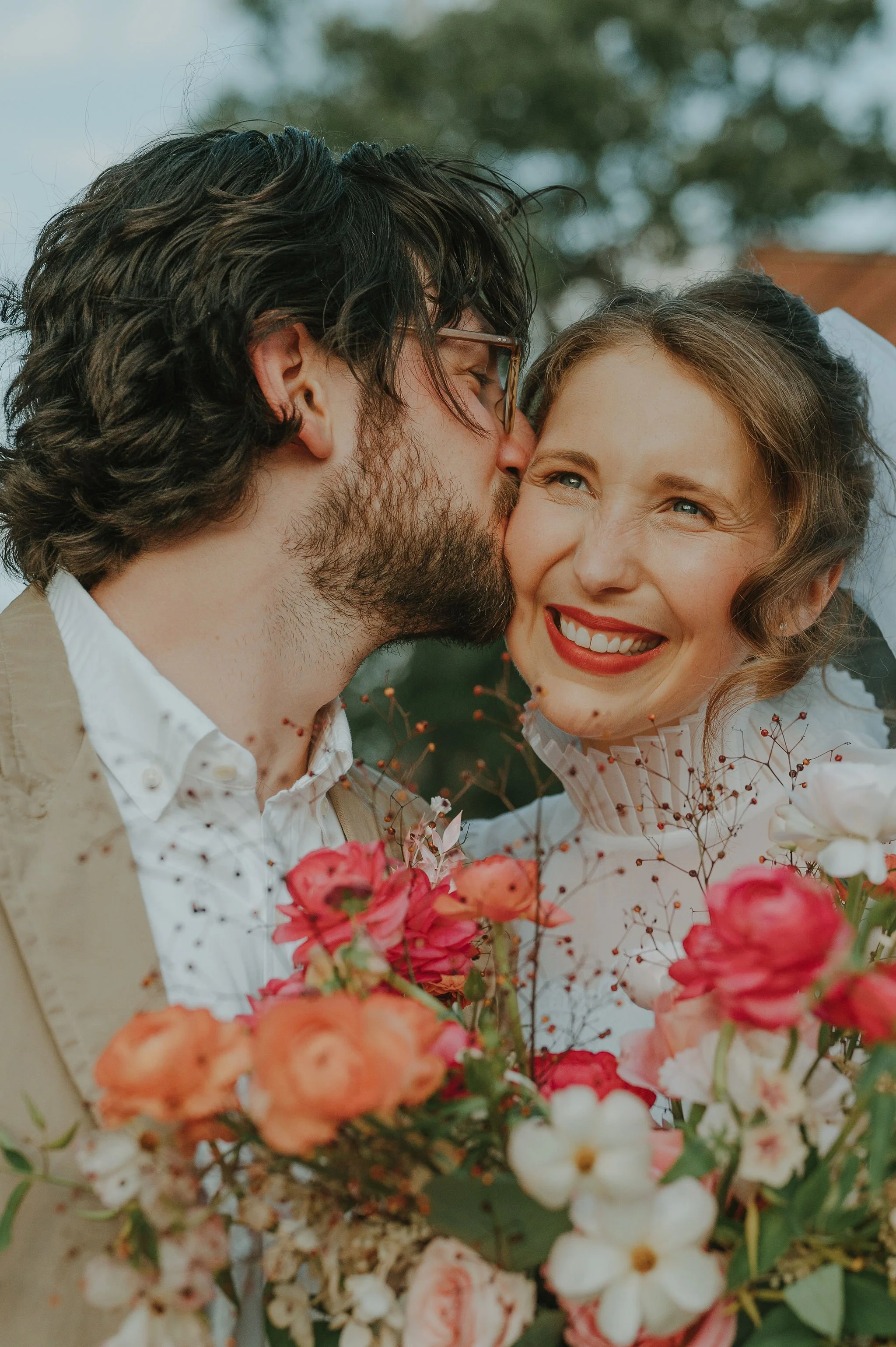 A man kisses a woman on her cheek during a floral outdoor event, with both smiling and surrounded by colorful flowers.