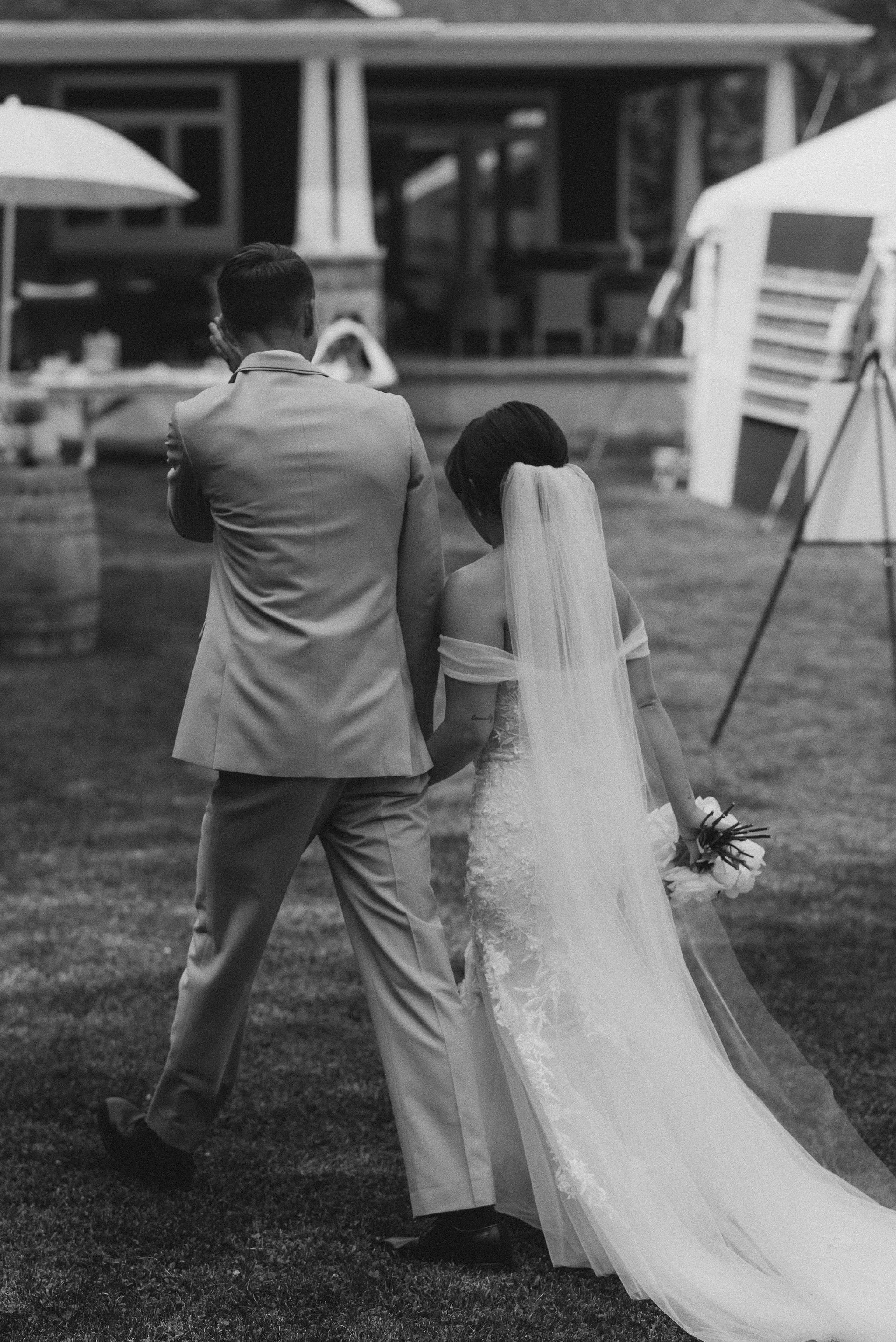 Black and white image of a bride and groom walking away, holding hands, during a wedding ceremony on a grassy outdoor area.