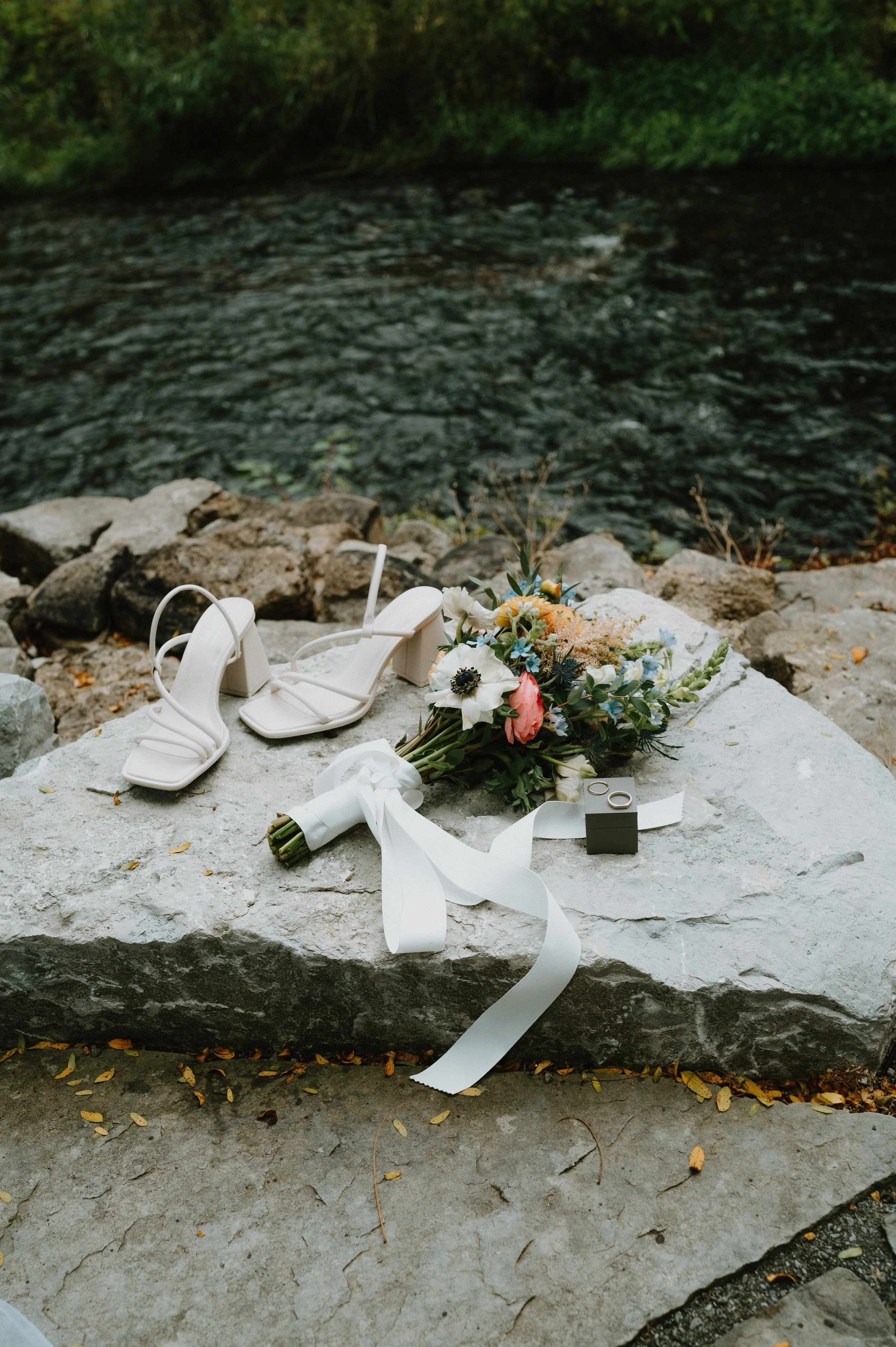 A white floral bouquet, a pair of white high-heeled sandals, and a small black box with rings on a large stone by a river.