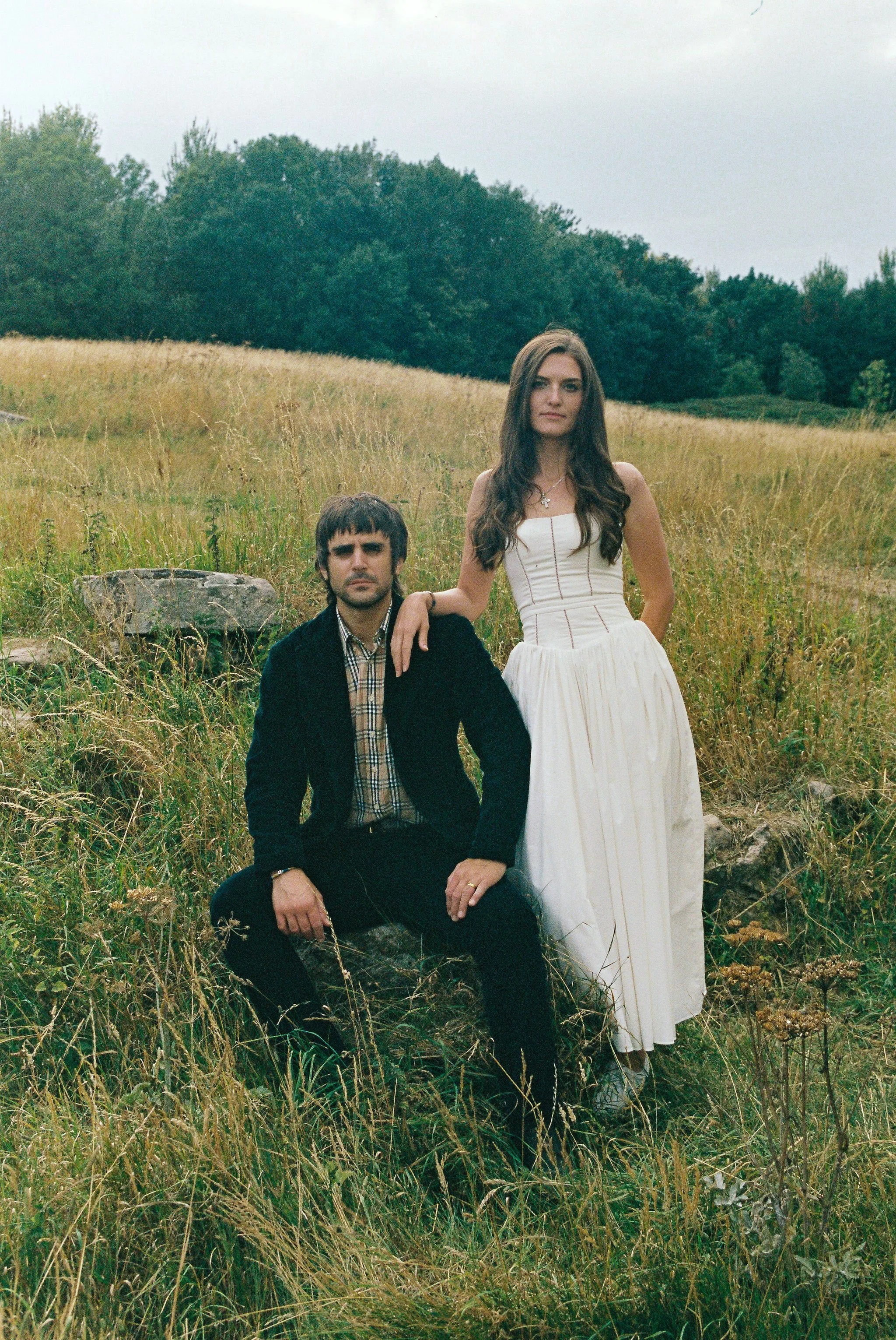 A man and woman in formal attire are in a grassy field with trees in the background. The woman is standing, and the man is sitting on a rock, with her hand resting on his shoulder.