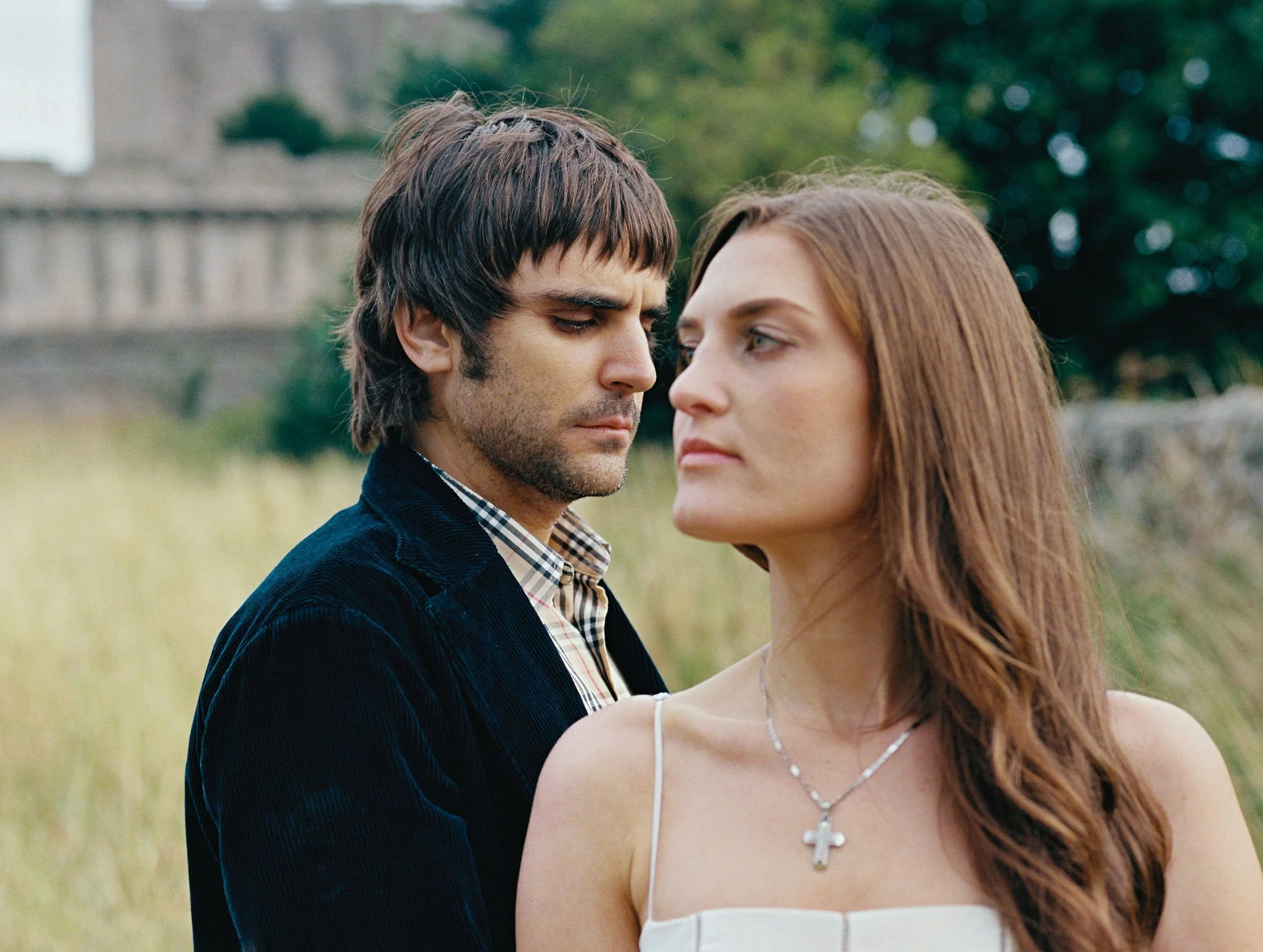 A man and woman standing outdoors with greenery in the background, facing each other with serious expressions.