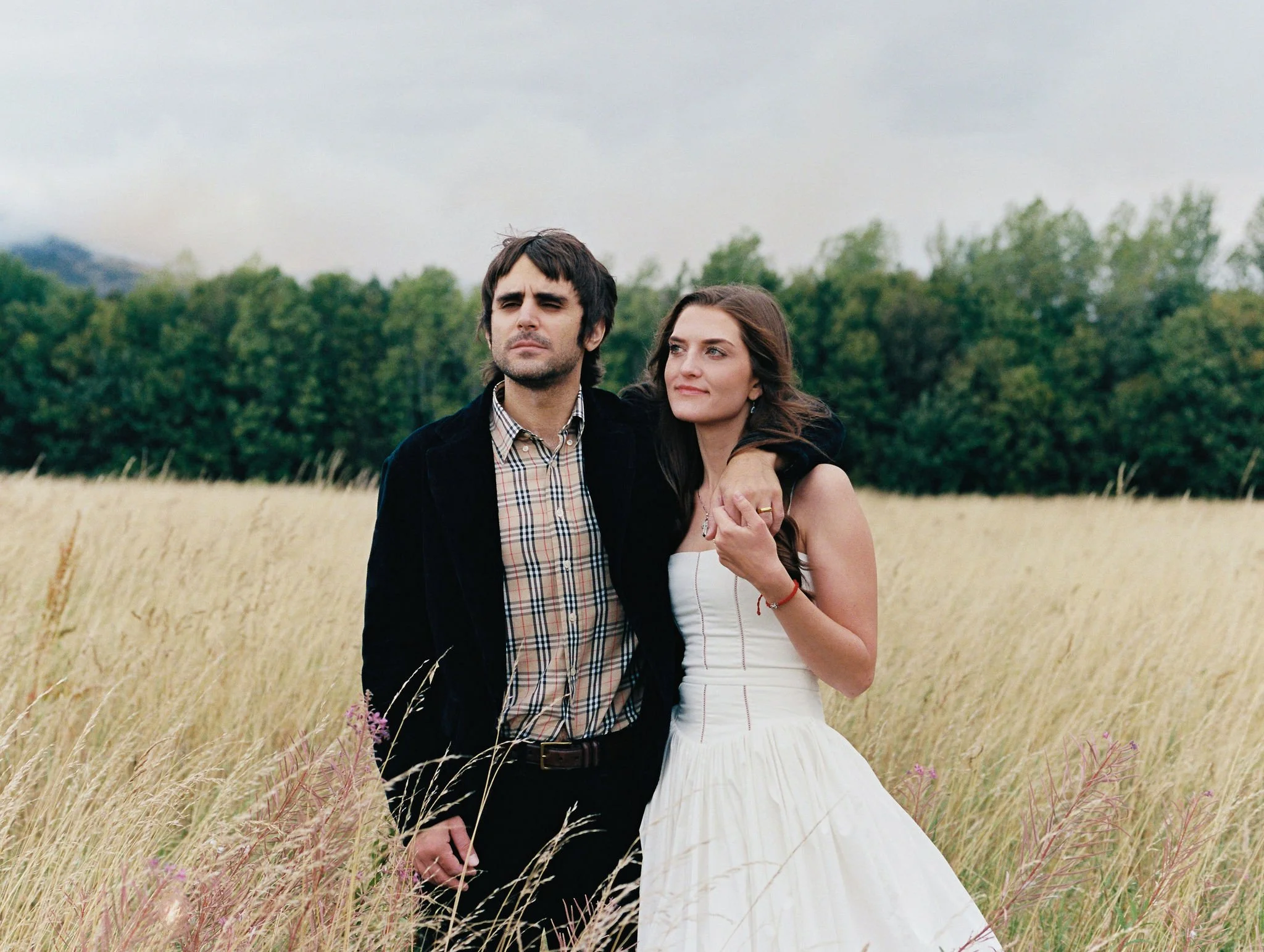 A man and woman stand together in a field of tall grass with a backdrop of green trees under a cloudy sky. The man has dark hair, a slight beard, and wears a black jacket over a plaid shirt. The woman has long dark hair and wears a white sleeveless d