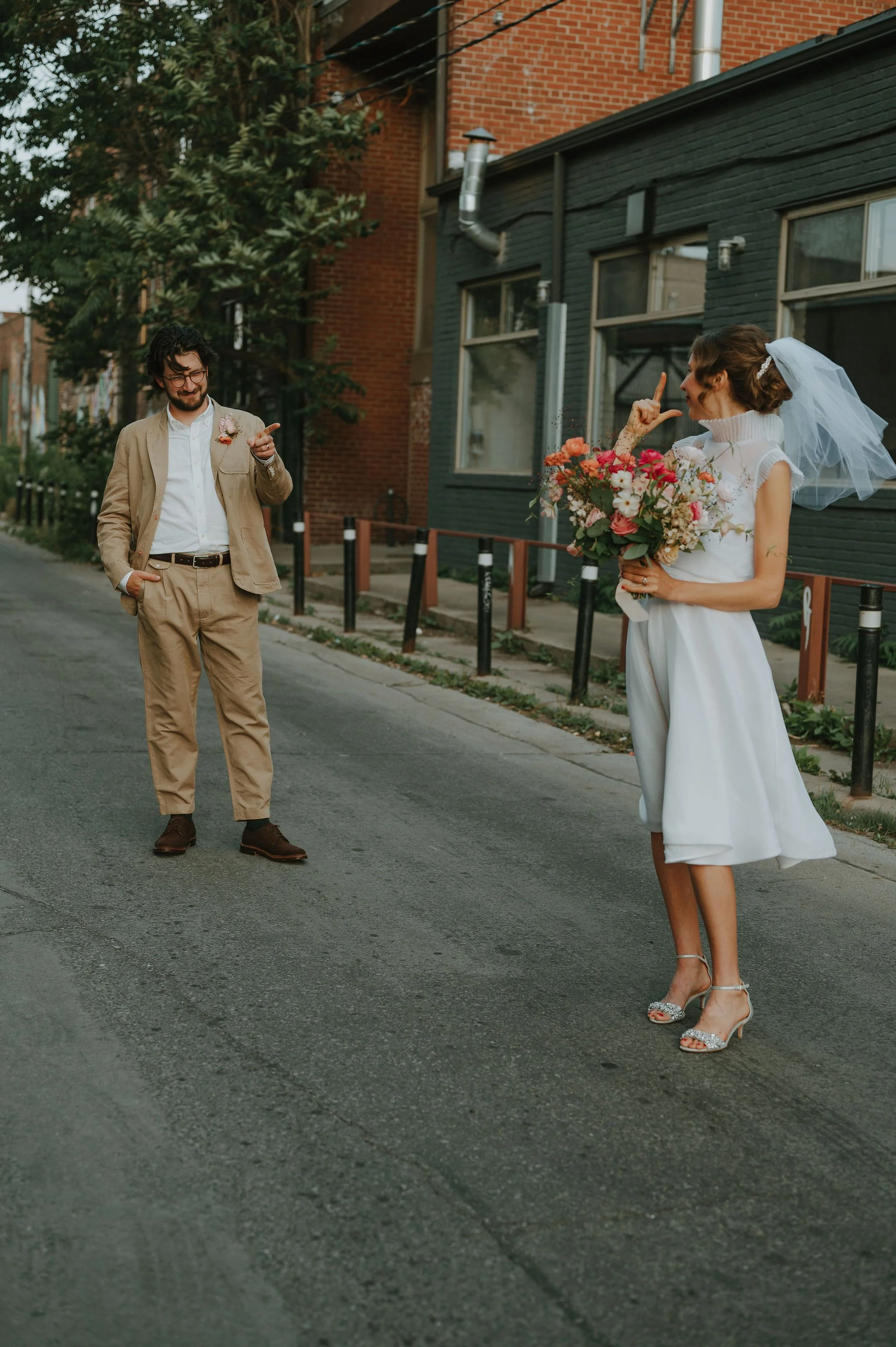 A bride in a white dress and veil holding a bouquet of flowers standing in an urban street, playfully pointing at a man in a beige suit with glasses who has one hand in his pocket, while the other points back at her.