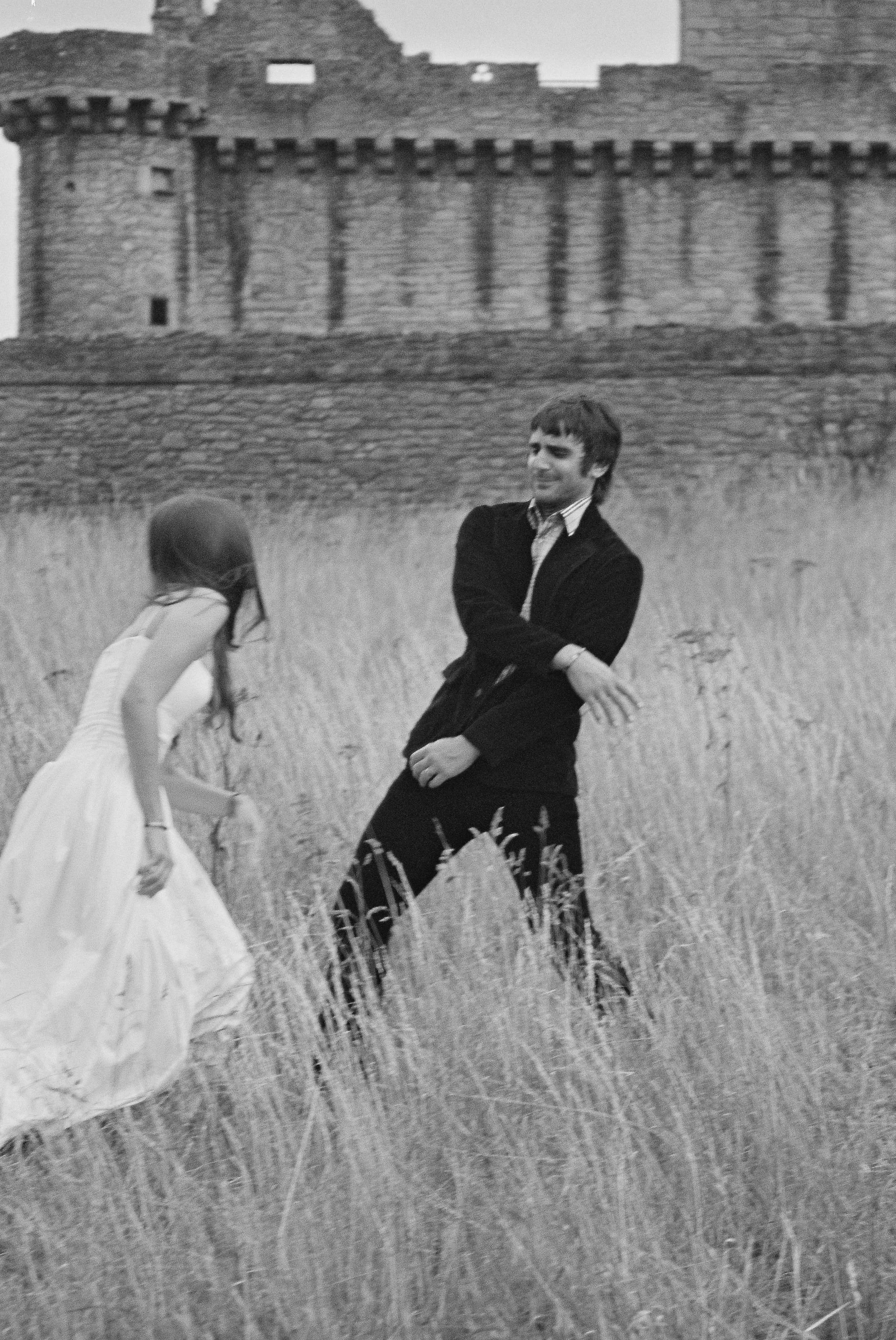 A young woman in a white dress and a young man in dark clothing dancing or playfully interacting in a grassy field, with a large stone castle or fortress in the background.