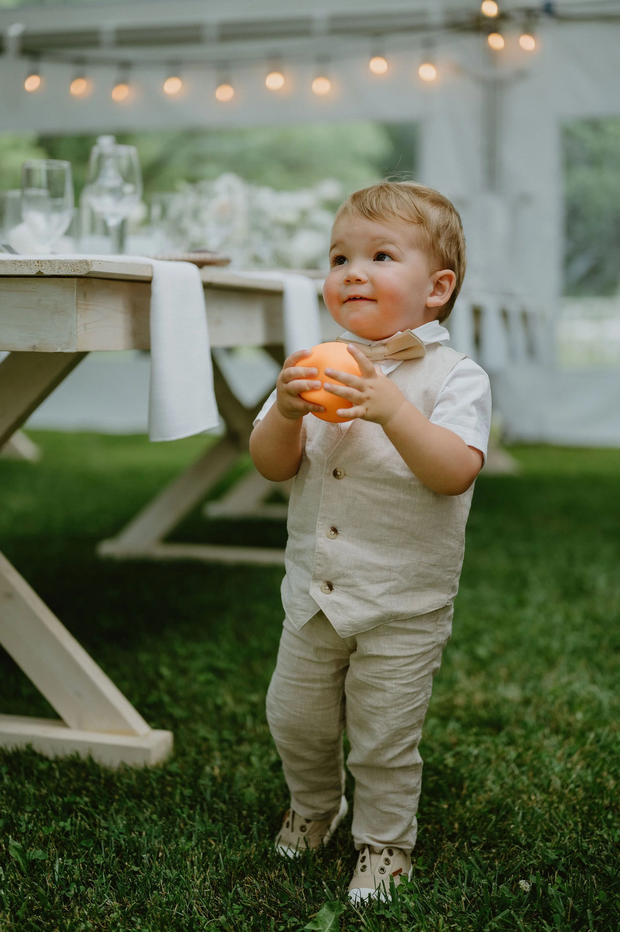 A young boy dressed in a beige vest, pants, and a bow tie stands outdoors on grass holding an orange ball, with a decorated table and string lights in the background.
