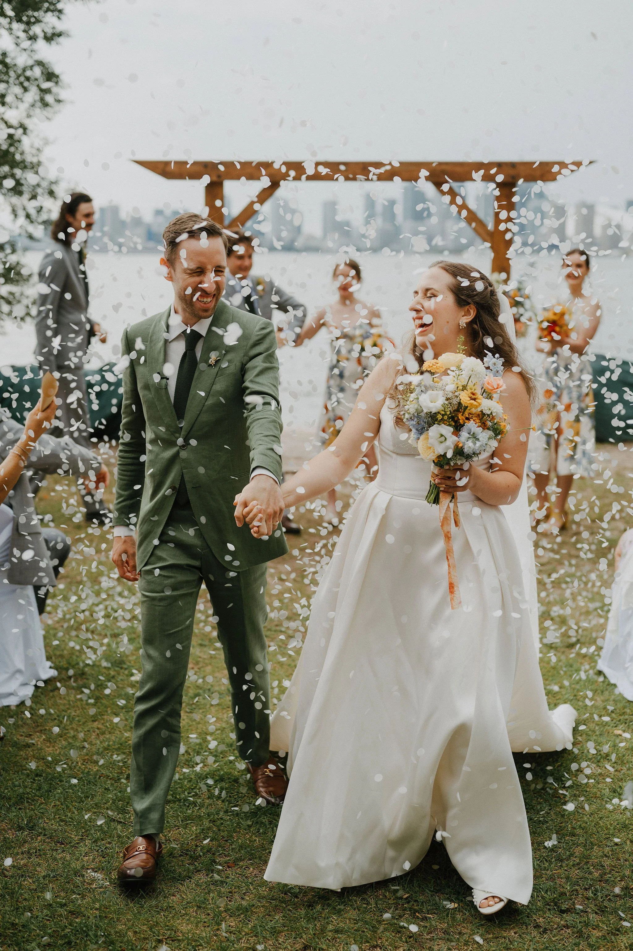 A newly married couple holding hands, smiling, and walking through falling confetti under a wooden arch at an outdoor wedding ceremony by the water, with bridesmaids and groomsmen in the background.