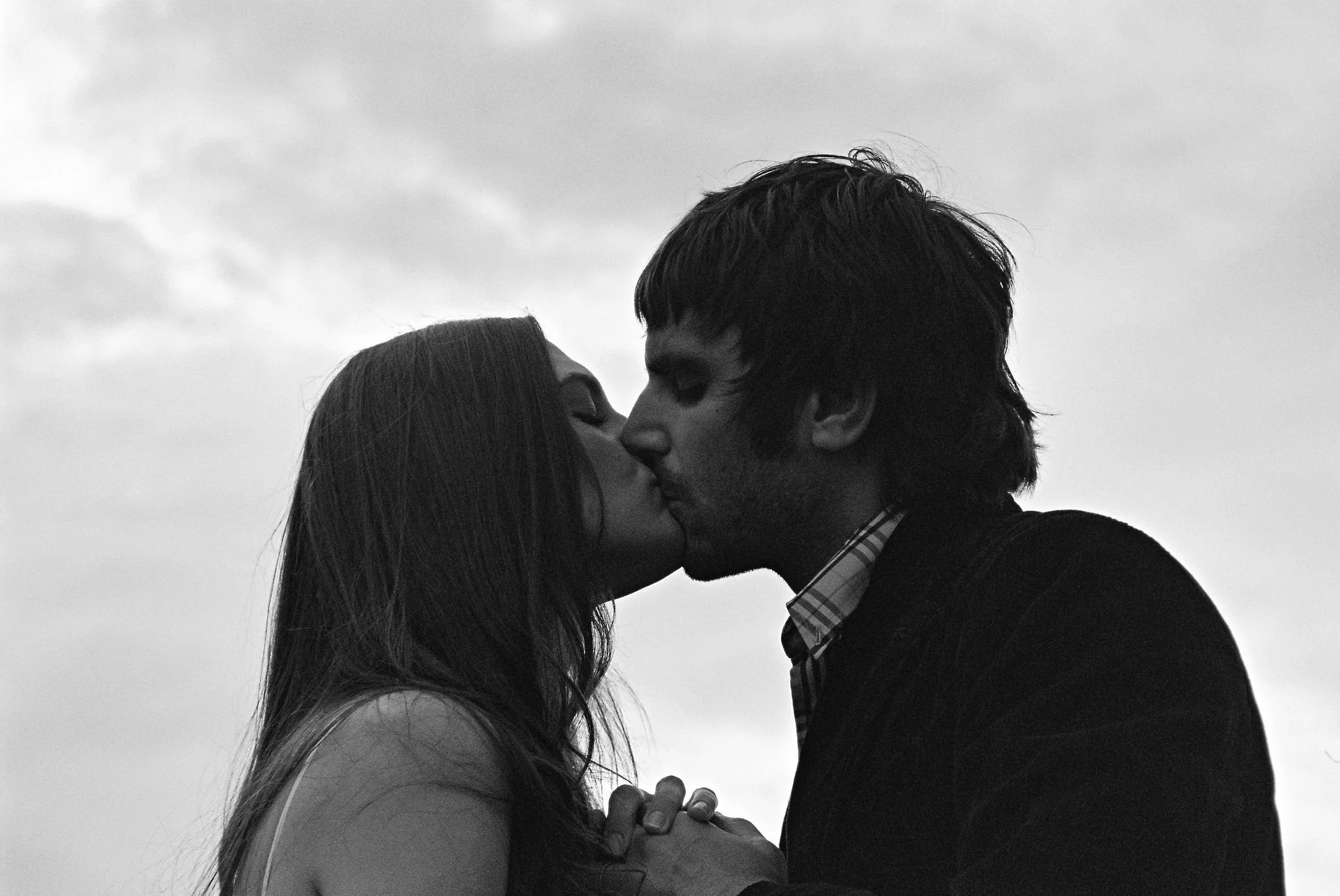 A black and white photo of a man and woman kissing outdoors, holding hands, against a cloudy sky.