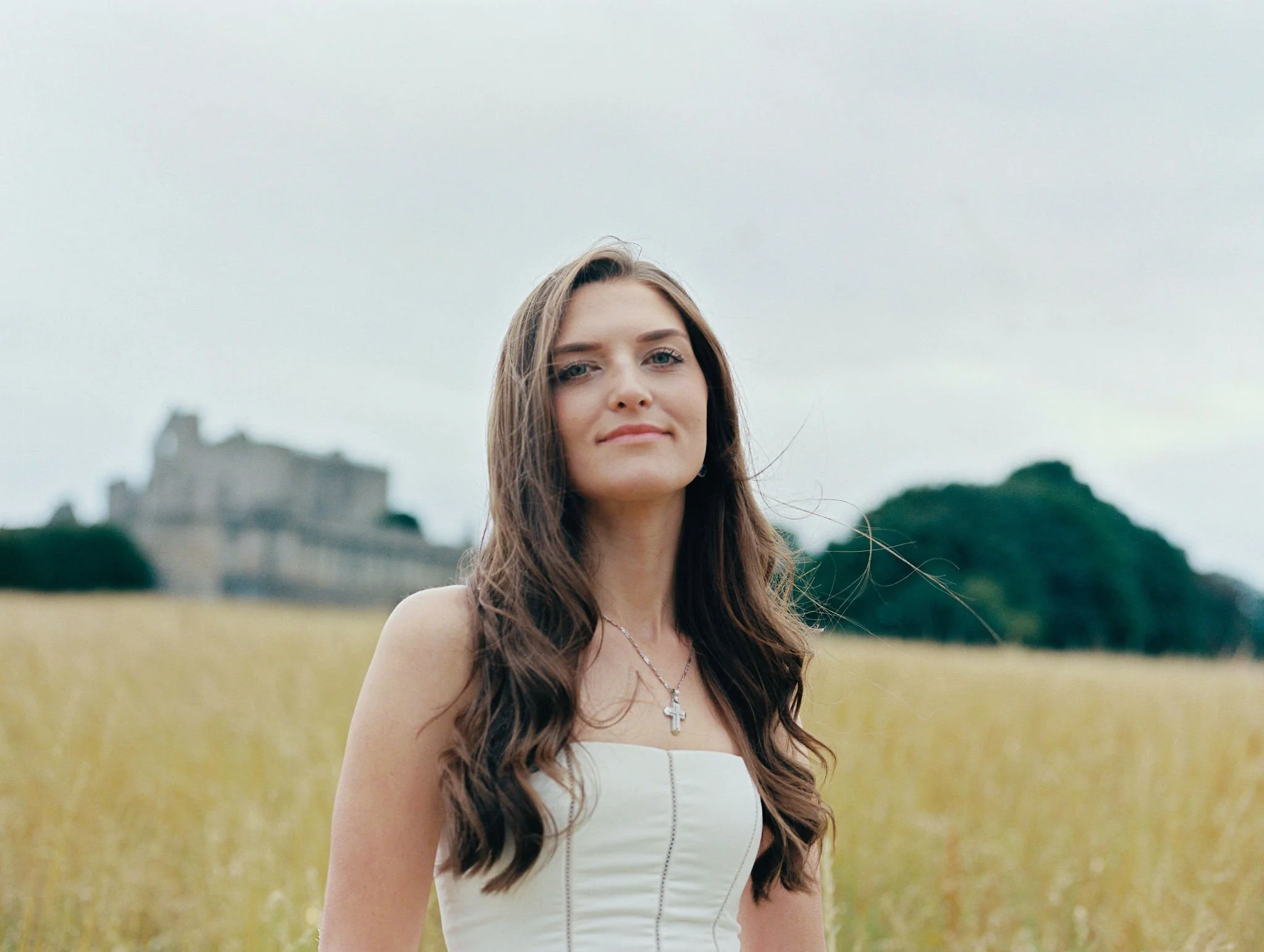 A woman with long brown hair standing outdoors in a field of yellow flowers, with a castle in the background under a cloudy sky.