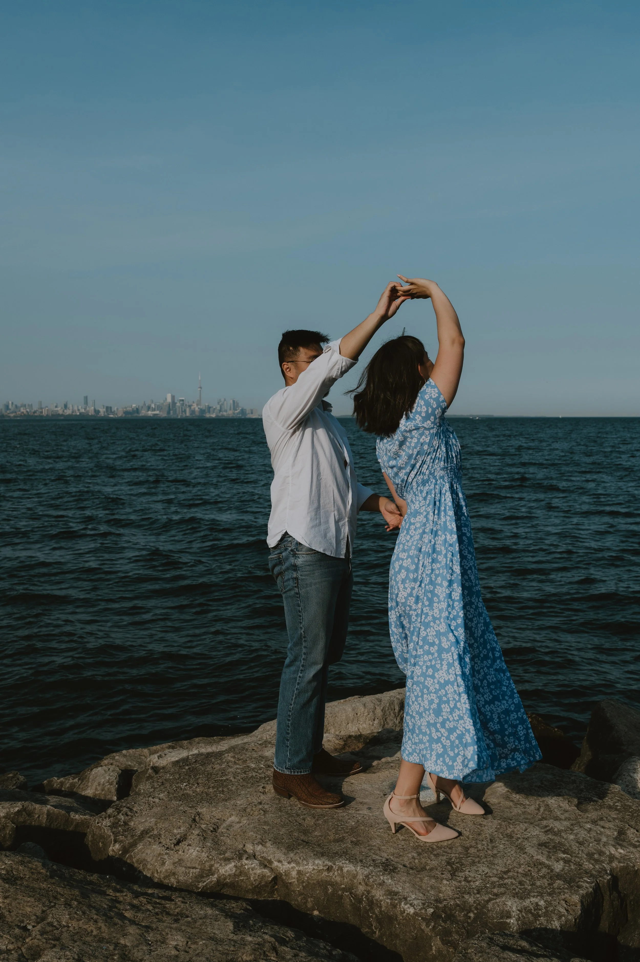 A couple dancing on rocks by the water with a city skyline in the background.