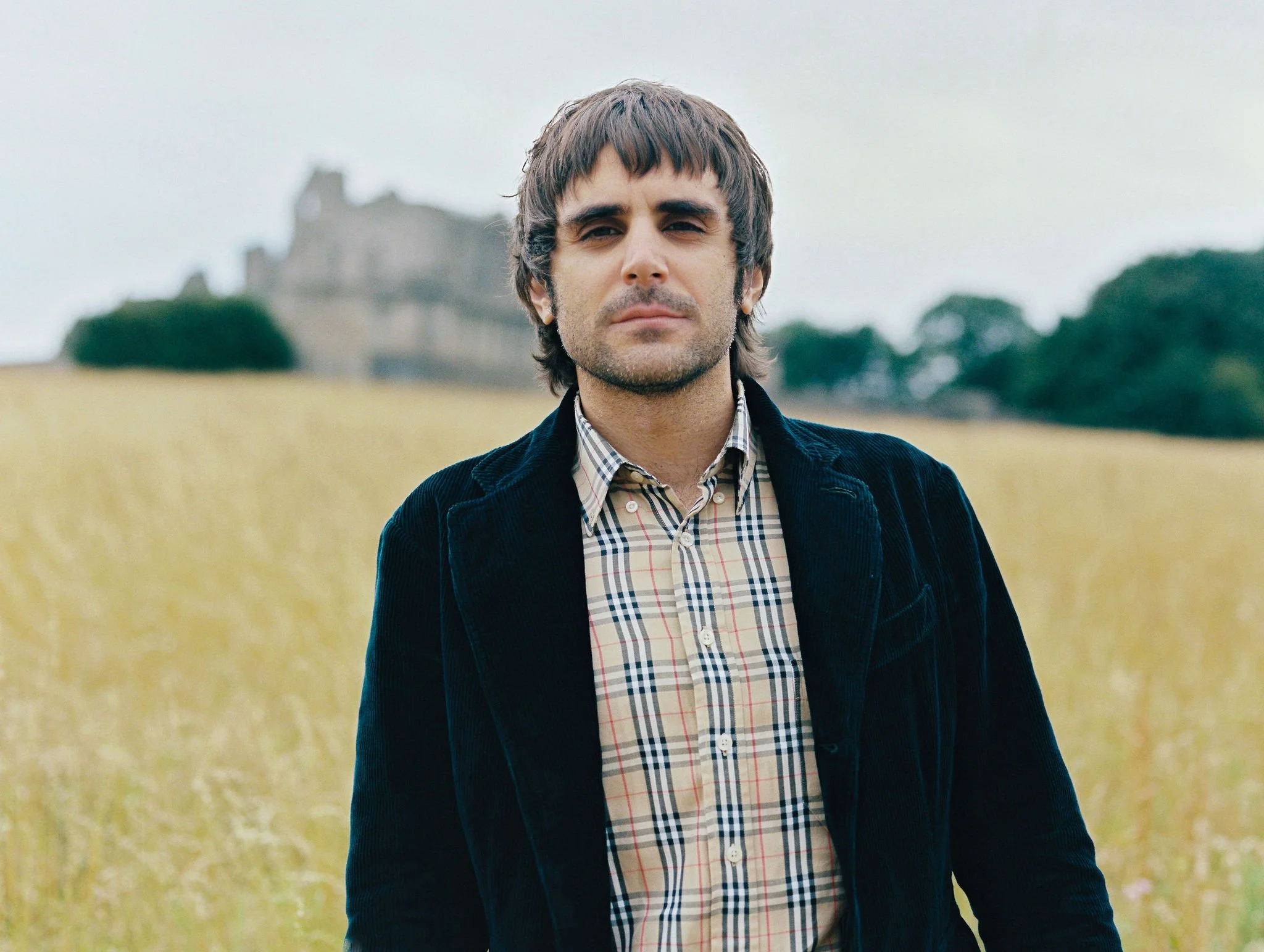 A man standing outdoors in a field with a castle in the background, wearing a plaid shirt and a dark jacket.