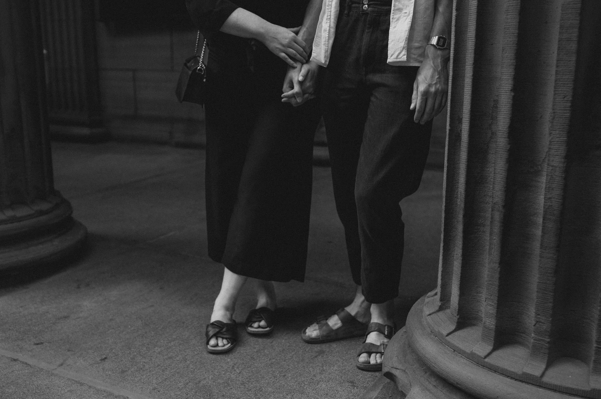 A close-up black-and-white photograph of two people holding hands, with their torsos and legs visible. They are standing near a column in an indoor setting, wearing casual clothing and sandals.