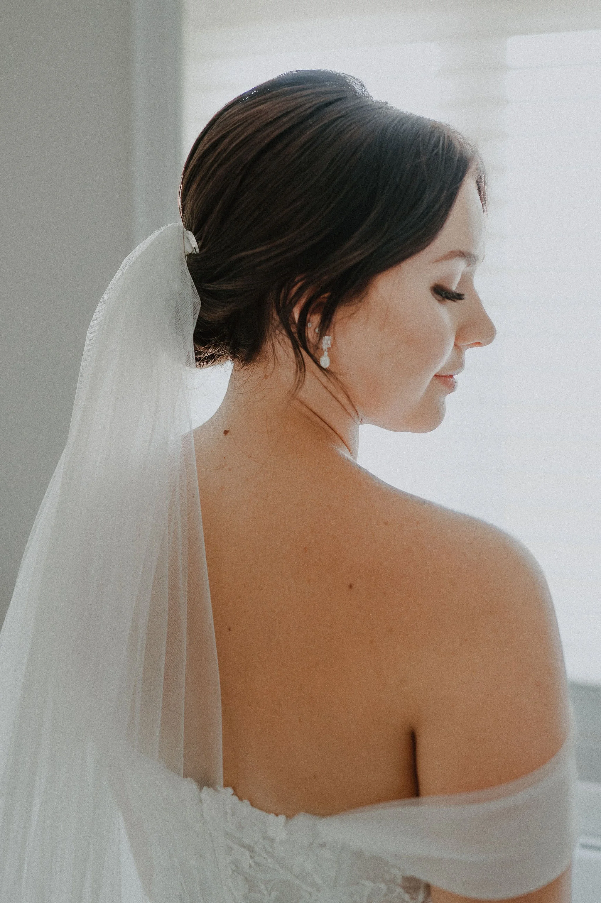 Bride with dark brown hair in an updo, wearing a veil and pearl earrings, looking down on her shoulder, soft natural light from a window in the background.