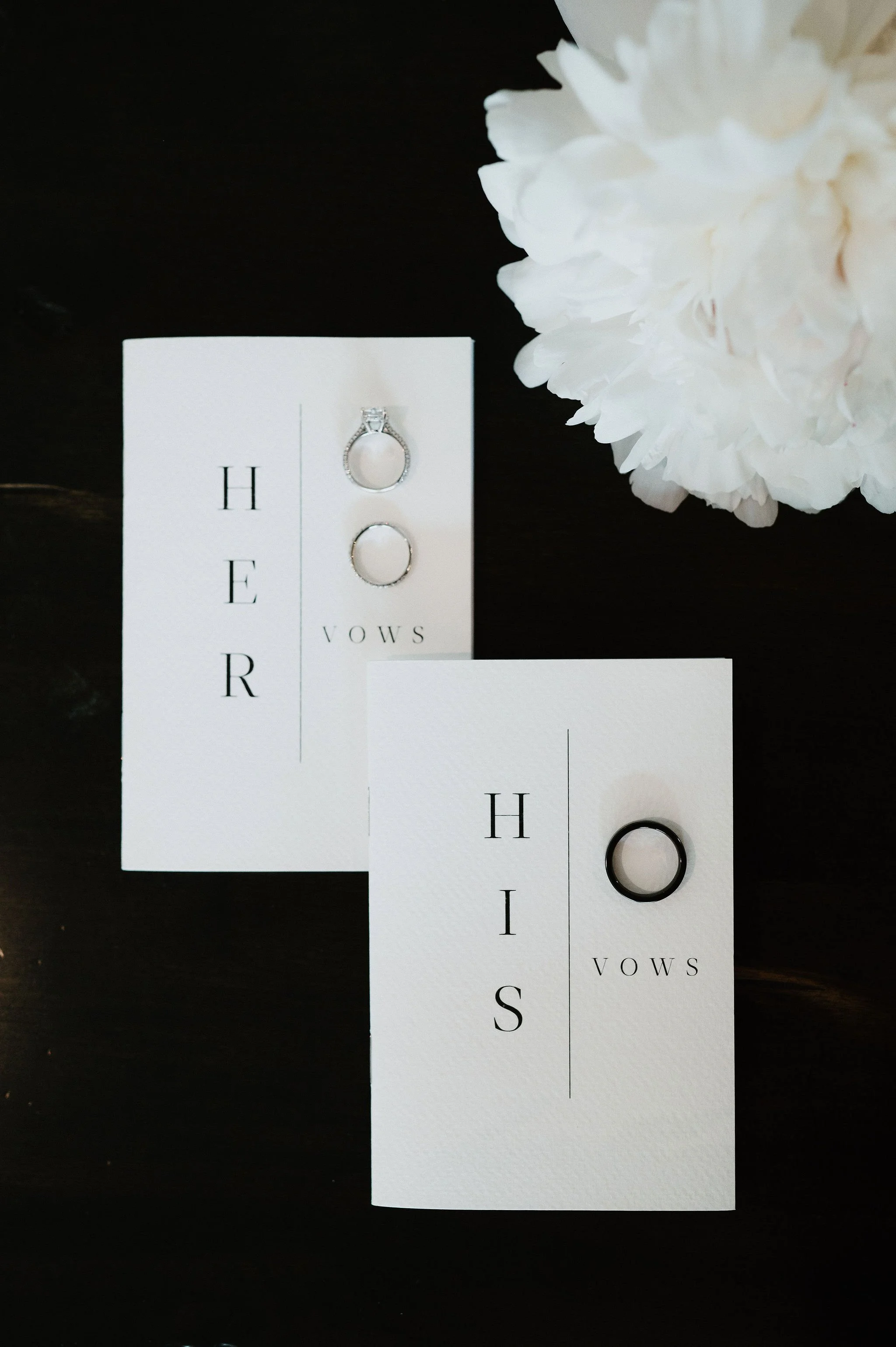 Wedding vows on white cards, with engagement and wedding rings, and white flower in the background.