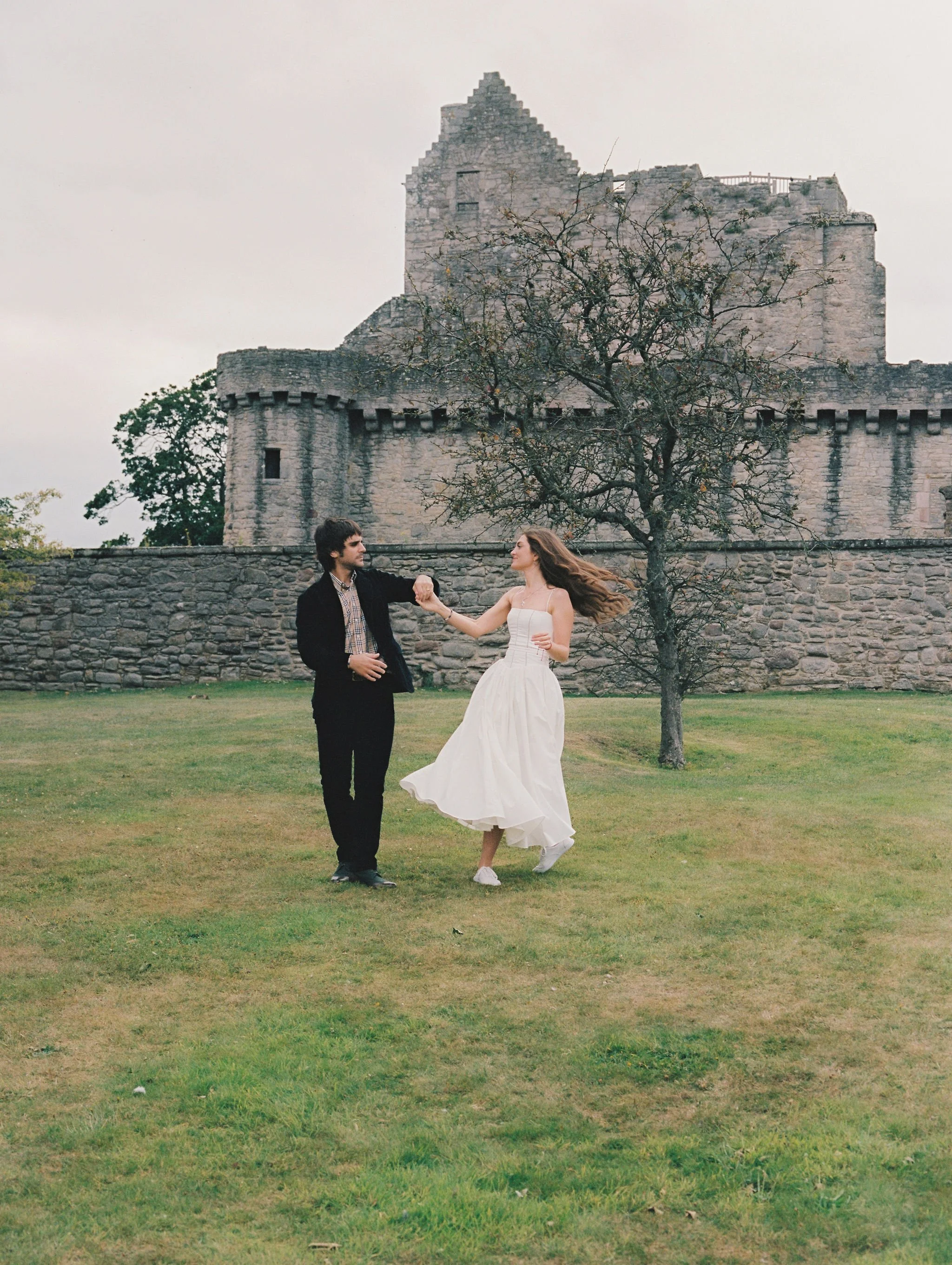 A couple dancing outdoors in front of a castle with a tree in the background.