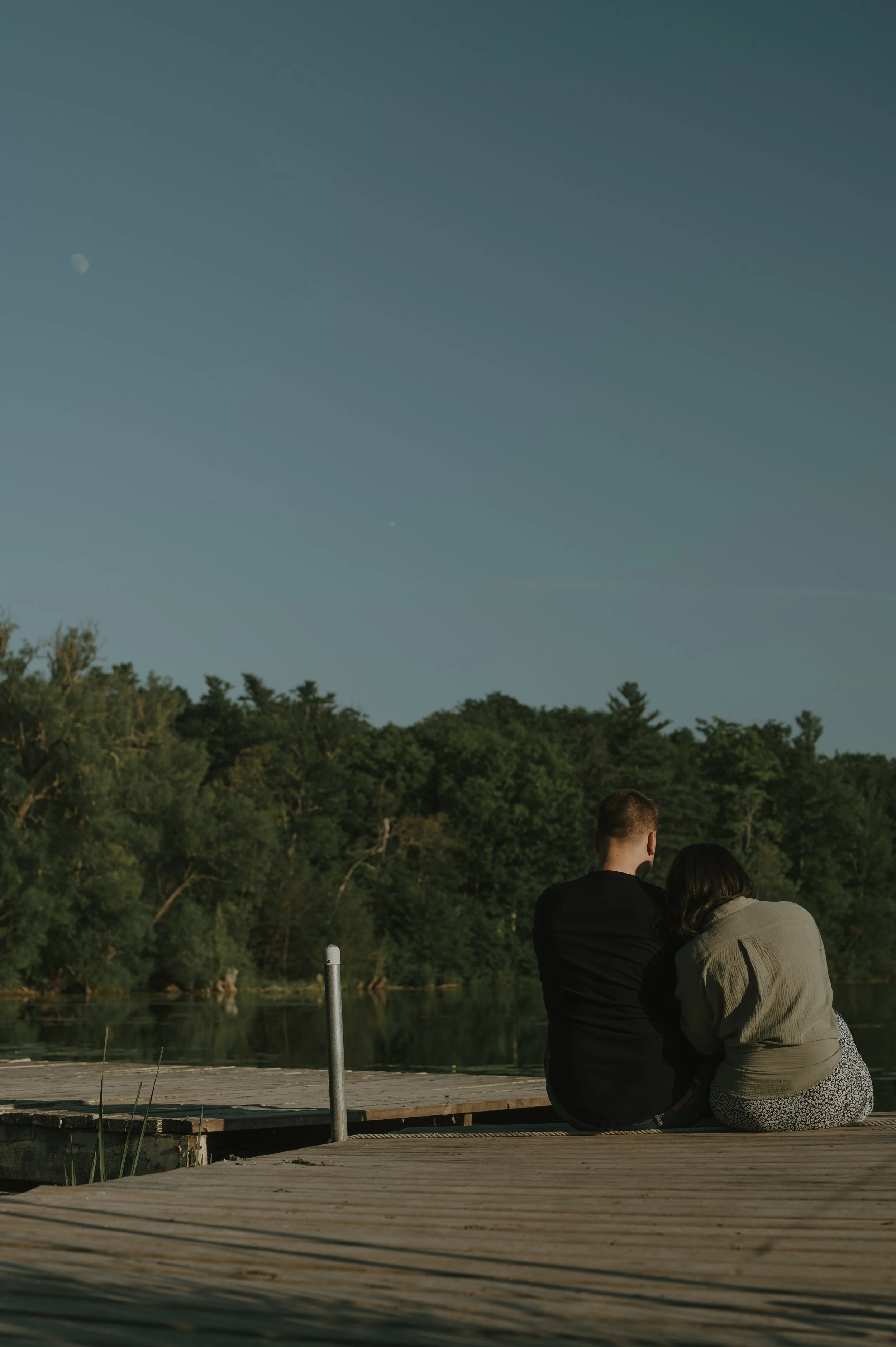 A couple sitting on a wooden dock by a lake, surrounded by green trees, with the moon visible in the early evening sky.