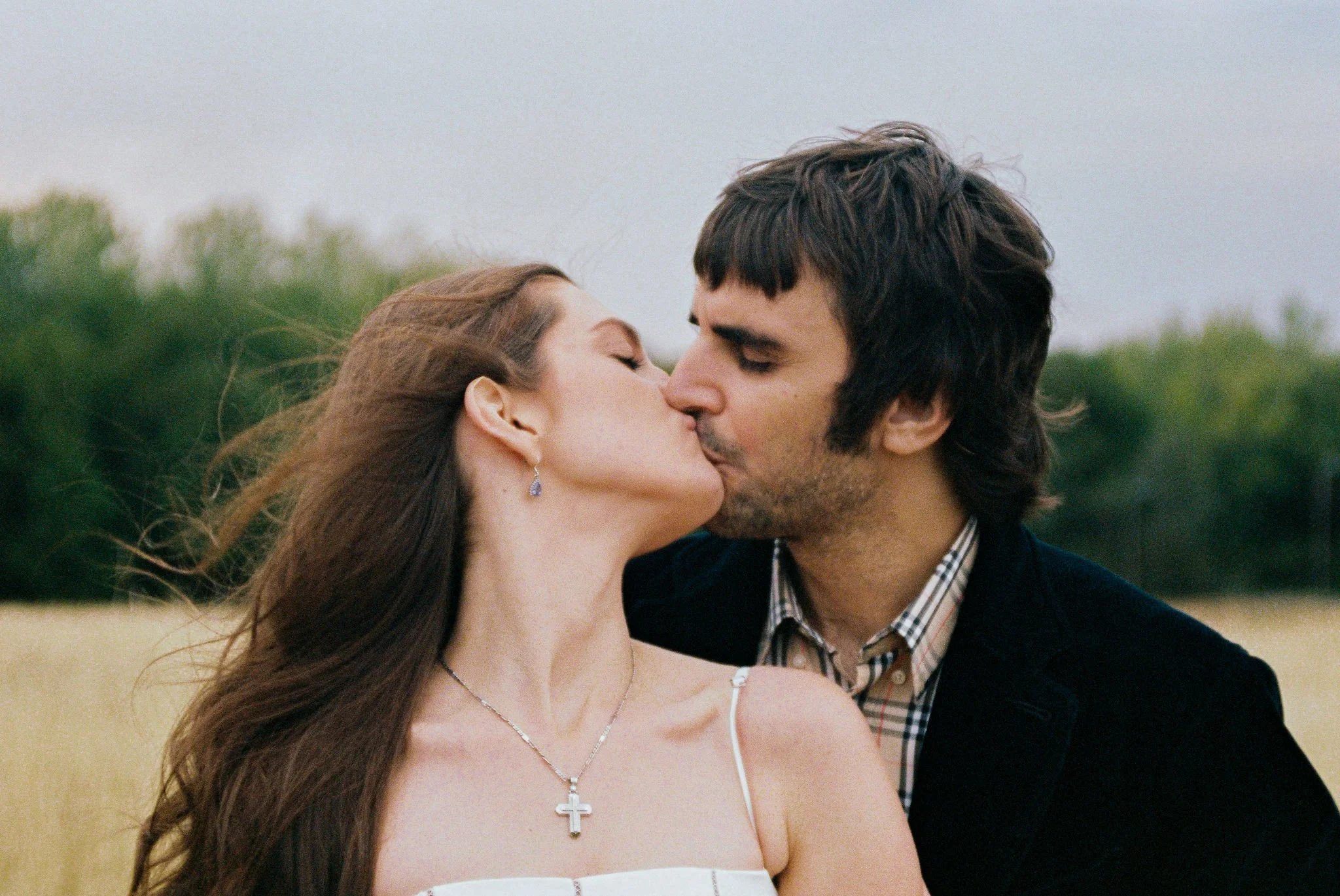 A man and woman kissing outdoors in a field with trees in the background.