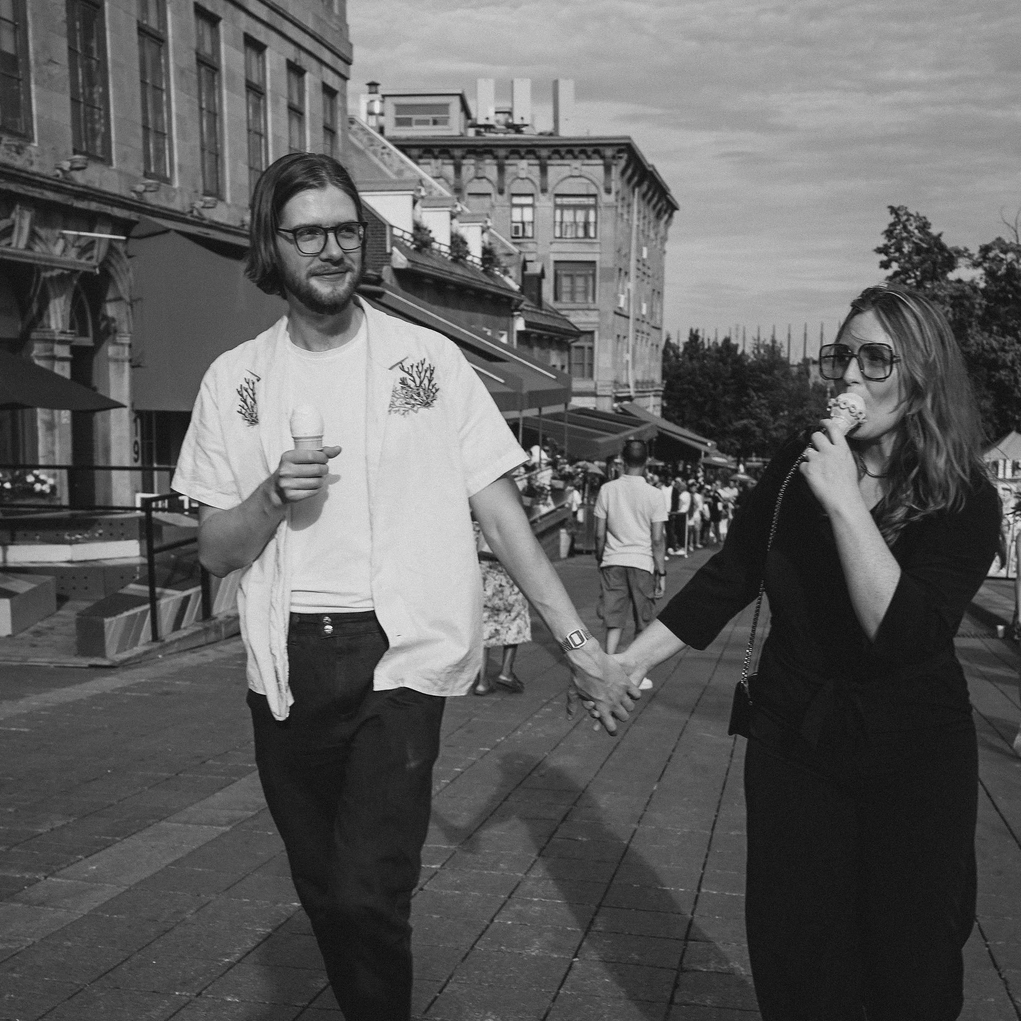 A black-and-white photo of a couple holding hands while walking outdoors on a city street, each eating ice cream cones, with buildings and people in the background.