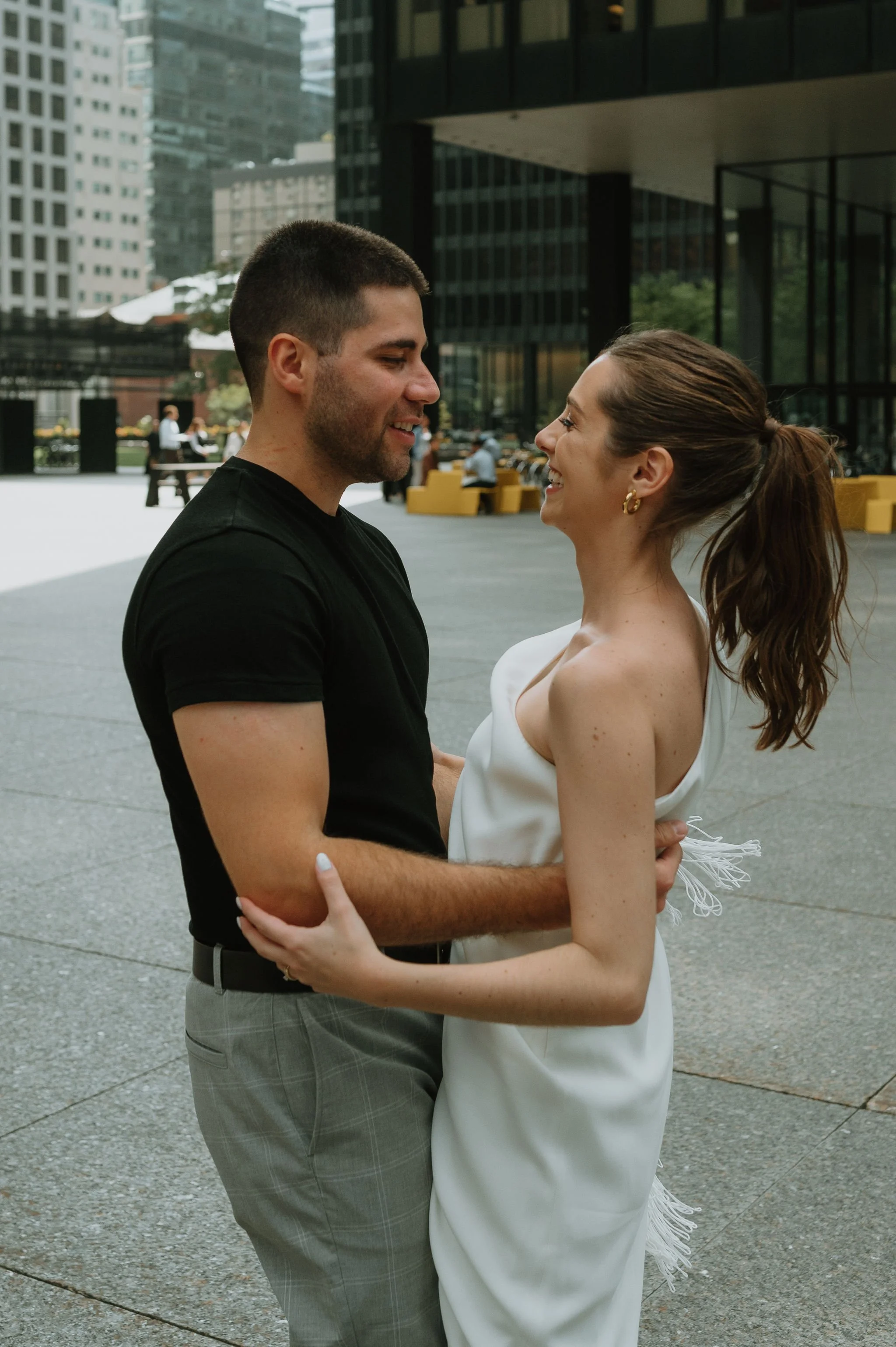 A couple smiling and holding each other outdoors in an urban setting, with tall buildings and a few people in the background.