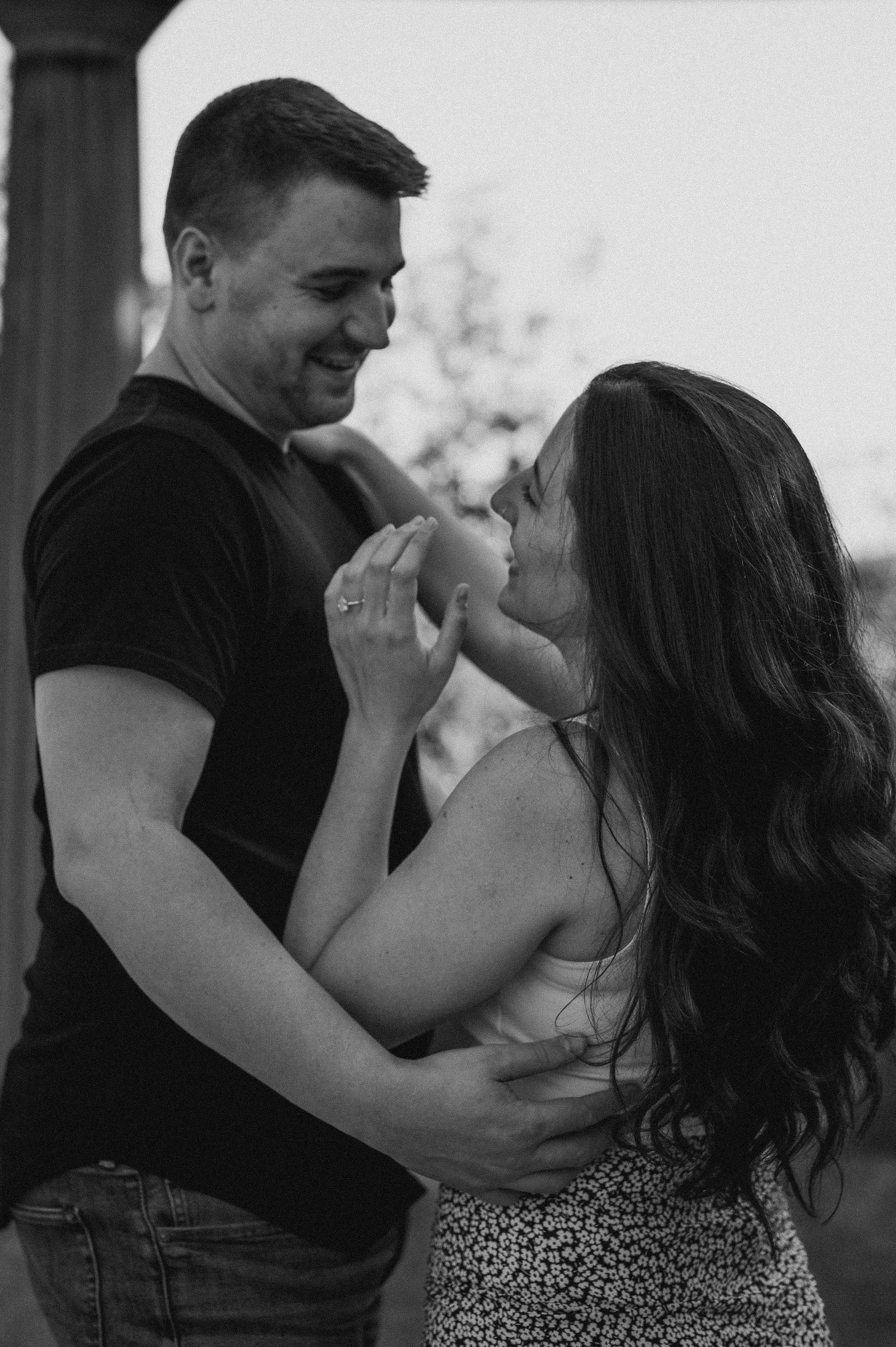 A black-and-white photo of a man and woman smiling at each other, dancing closely outside. The woman has long dark hair and is wearing a sleeveless top and patterned skirt. The man has short hair and is wearing a T-shirt and jeans.