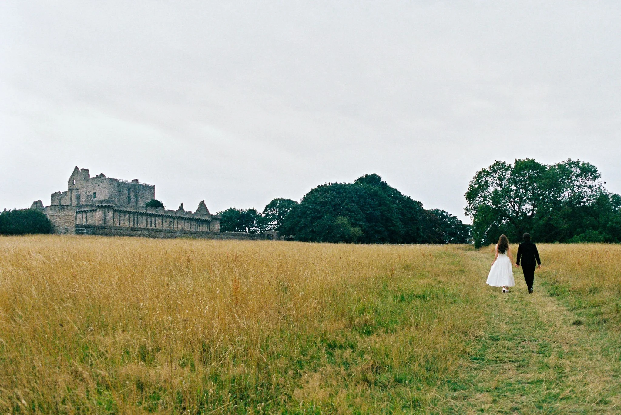 A bride and groom walking hand in hand along a grassy path through a field of tall golden grass, with a historic stone castle in the background, on a cloudy day.