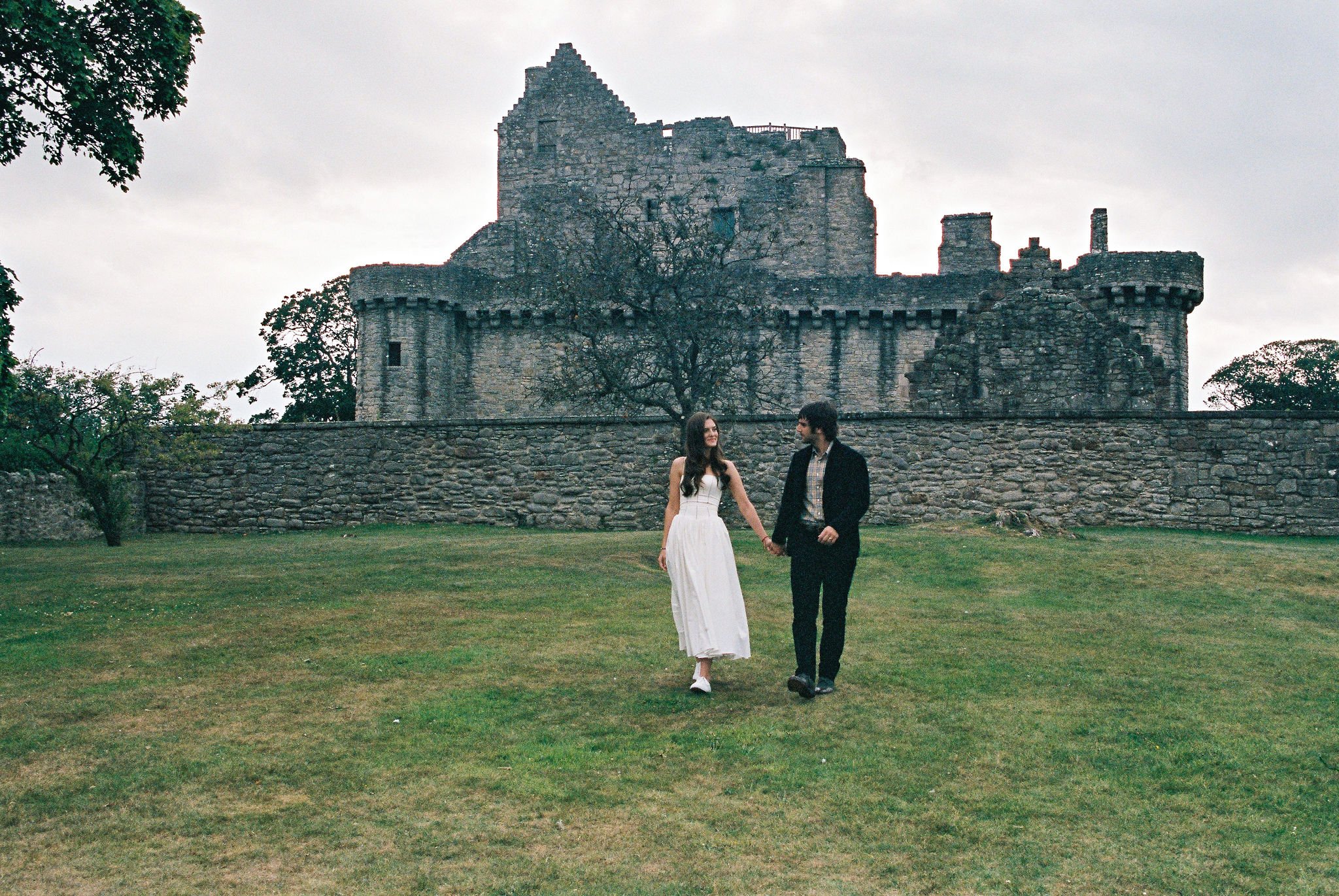 A couple walking hand in hand on a grassy field in front of an old stone castle surrounded by trees, overcast sky.