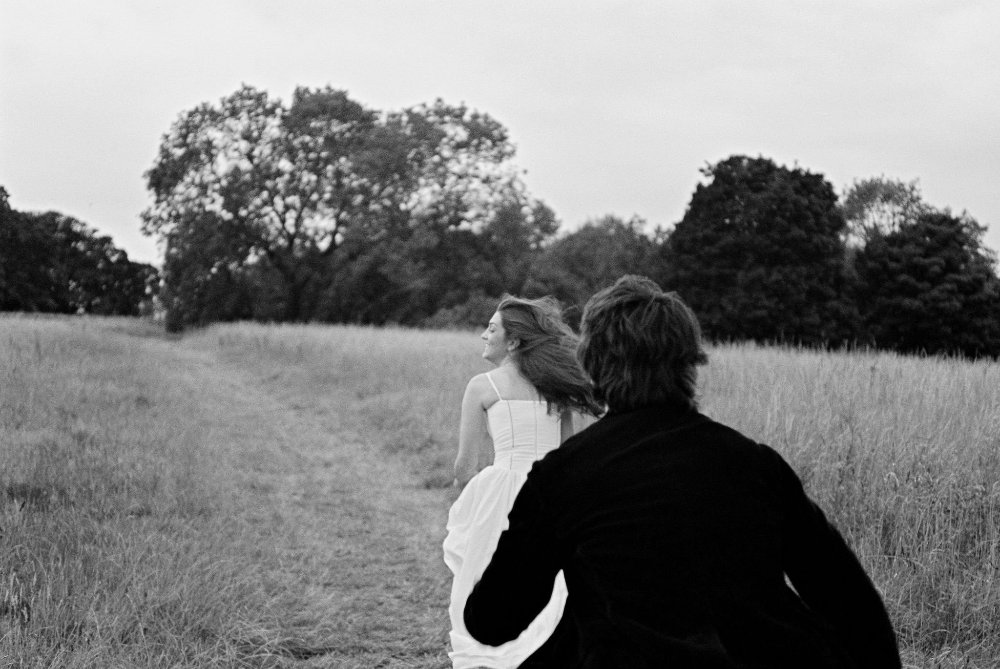 A black-and-white photo of a woman in a white dress walking on a grassy field, with a man in dark clothing in the foreground facing away, both surrounded by trees.