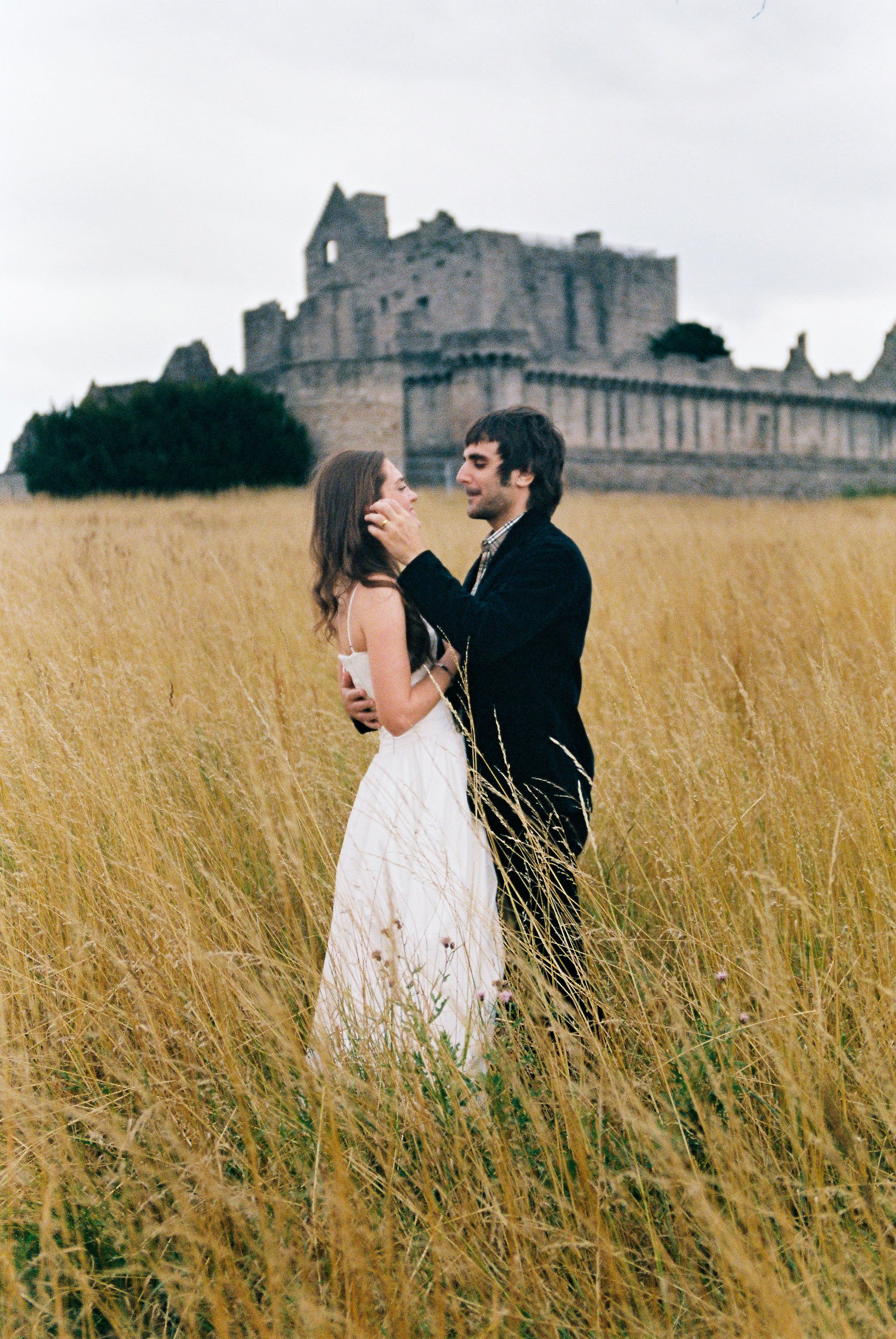 A romantic couple embracing in a field of tall, golden grass with an ancient castle in the background, under a cloudy sky.