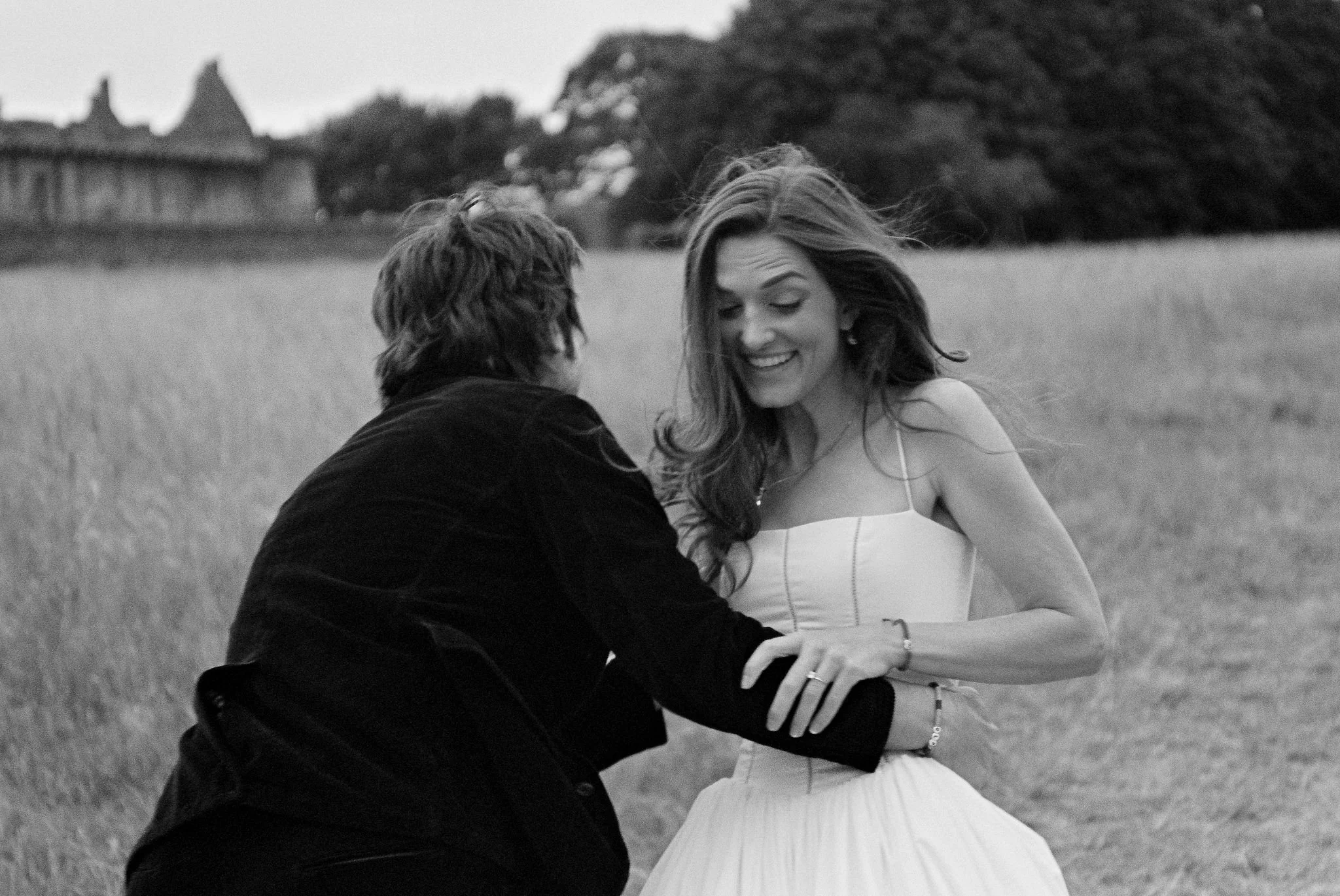A black-and-white photo of a woman in a white dress and a man in a dark shirt outdoors in a field, sharing a joyful moment.