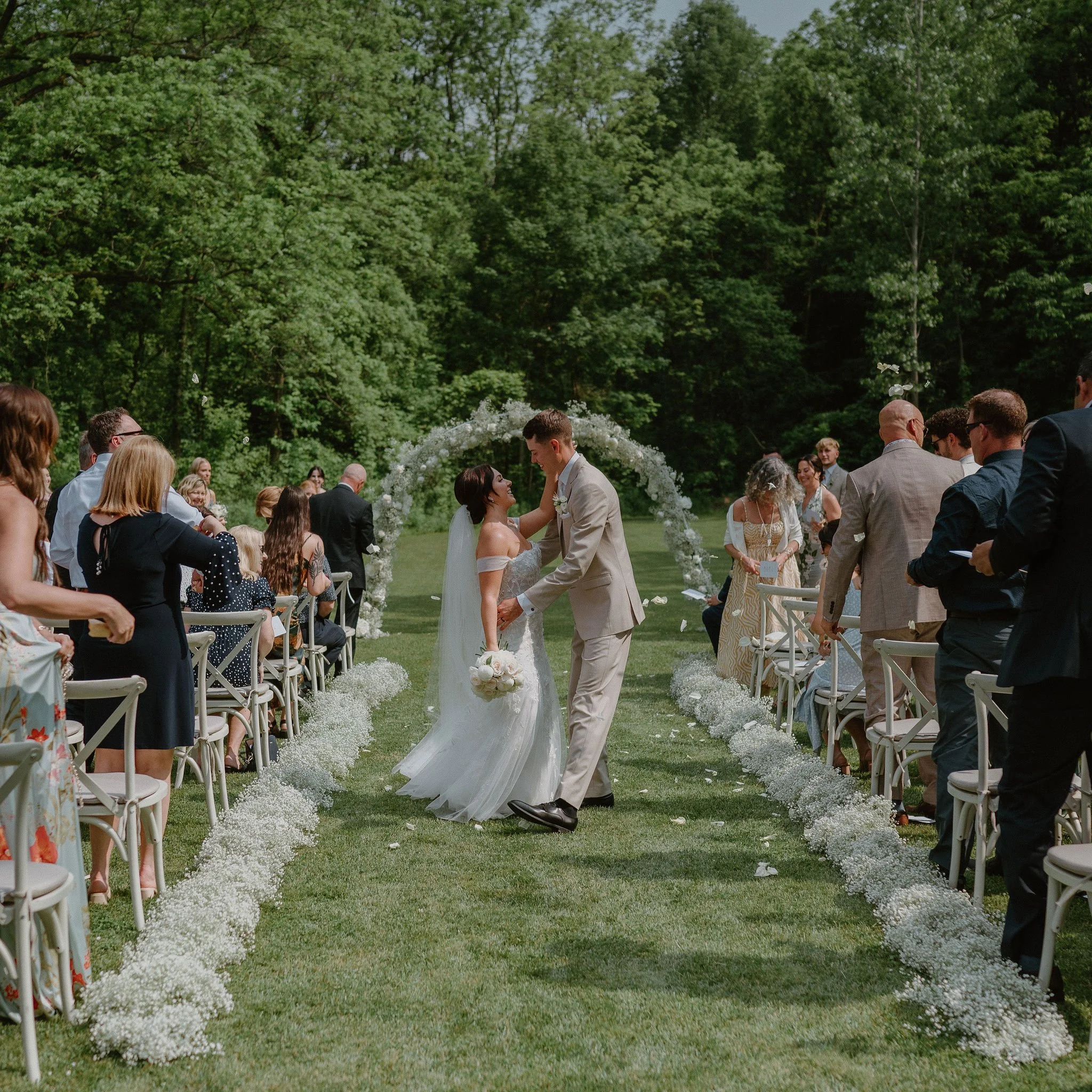 A bride and groom dancing during their outdoor wedding ceremony with guests seated on either side, and a floral arch in the background.