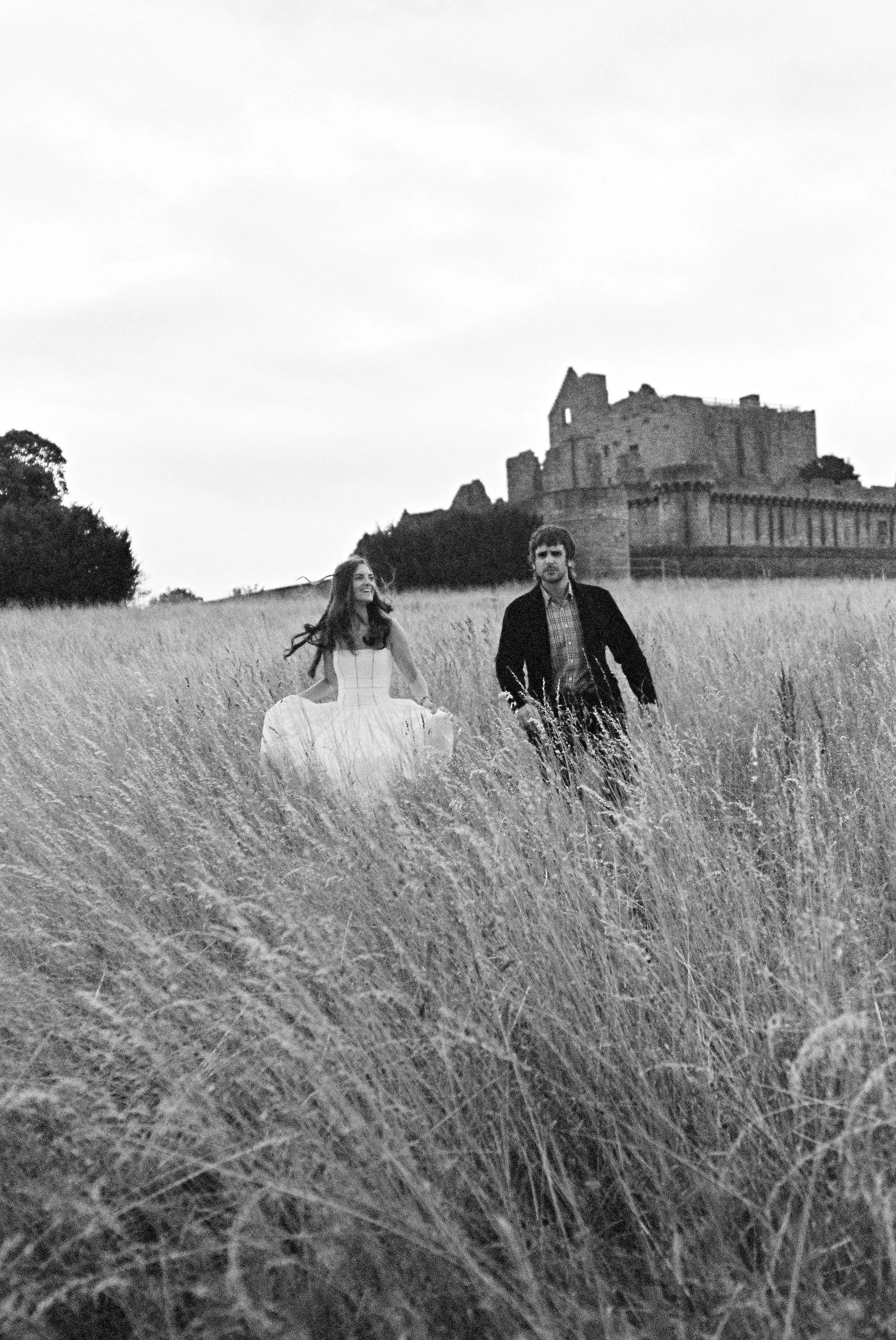 A black-and-white photo of a woman in a dress and a man in a jacket walking through a field of tall grass with a castle in the background.