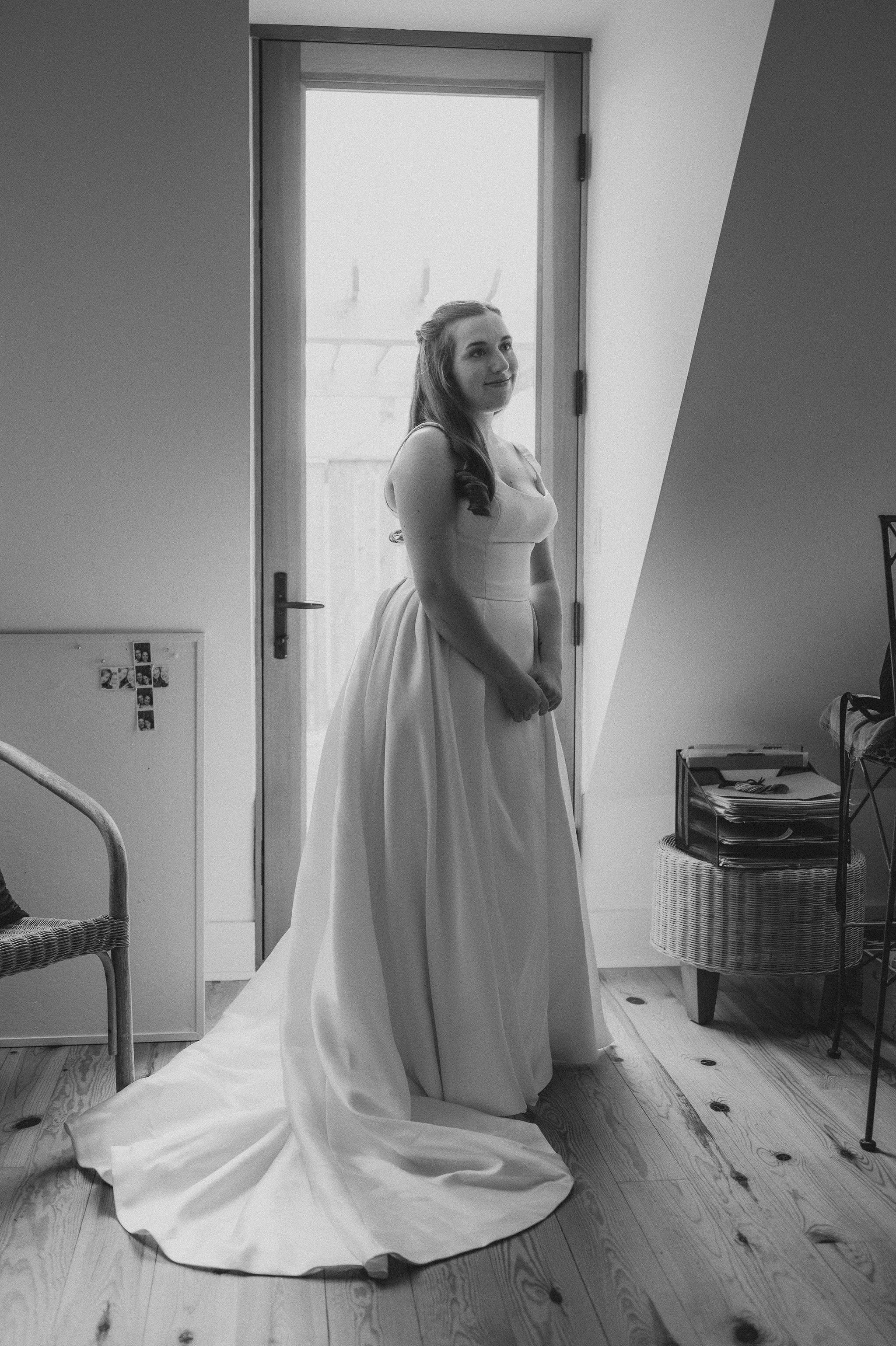A woman in a wedding dress standing indoors near a door, with a serene expression.