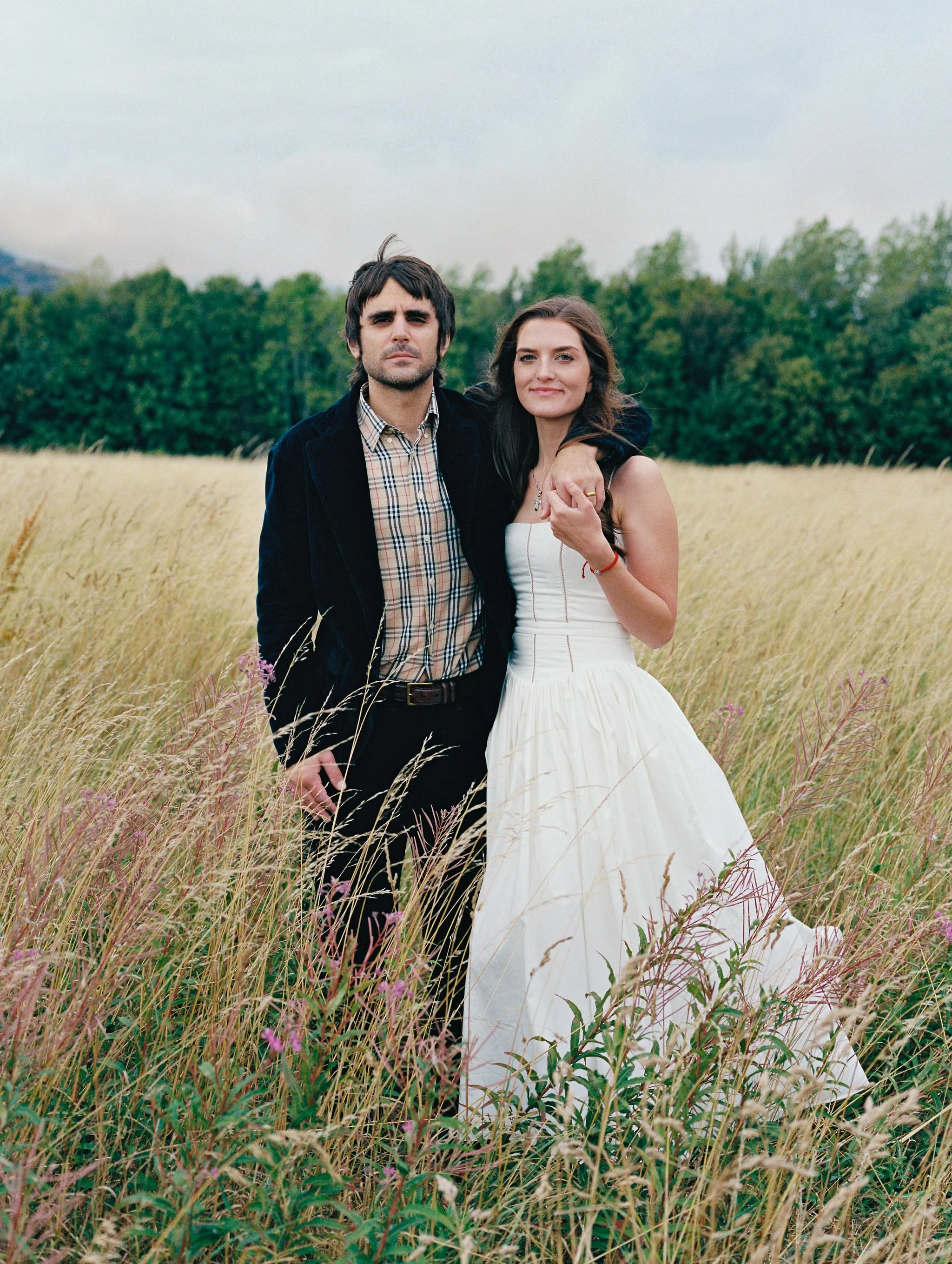 A couple standing close in a field of tall grass with trees in the background, the woman wearing a white dress and the man wearing a dark jacket and plaid shirt.