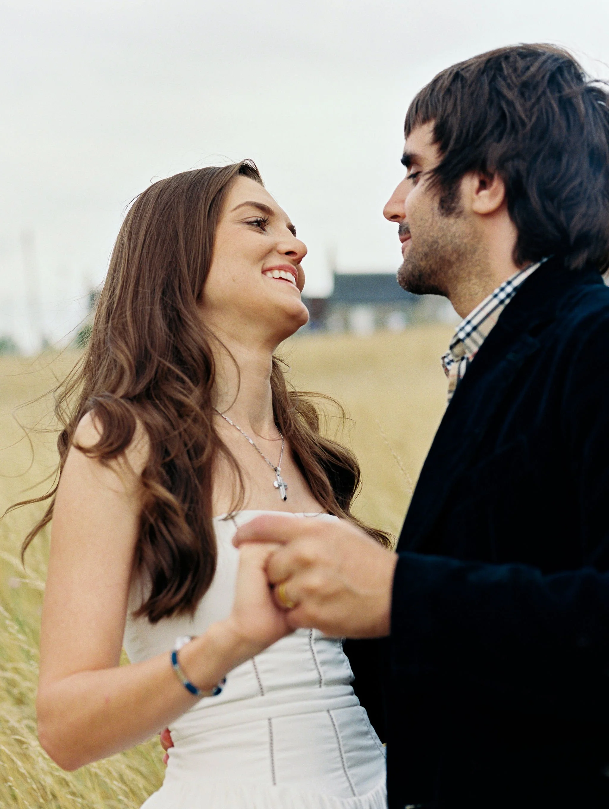 A young woman and a young man are holding hands and looking at each other in an outdoor field, smiling.