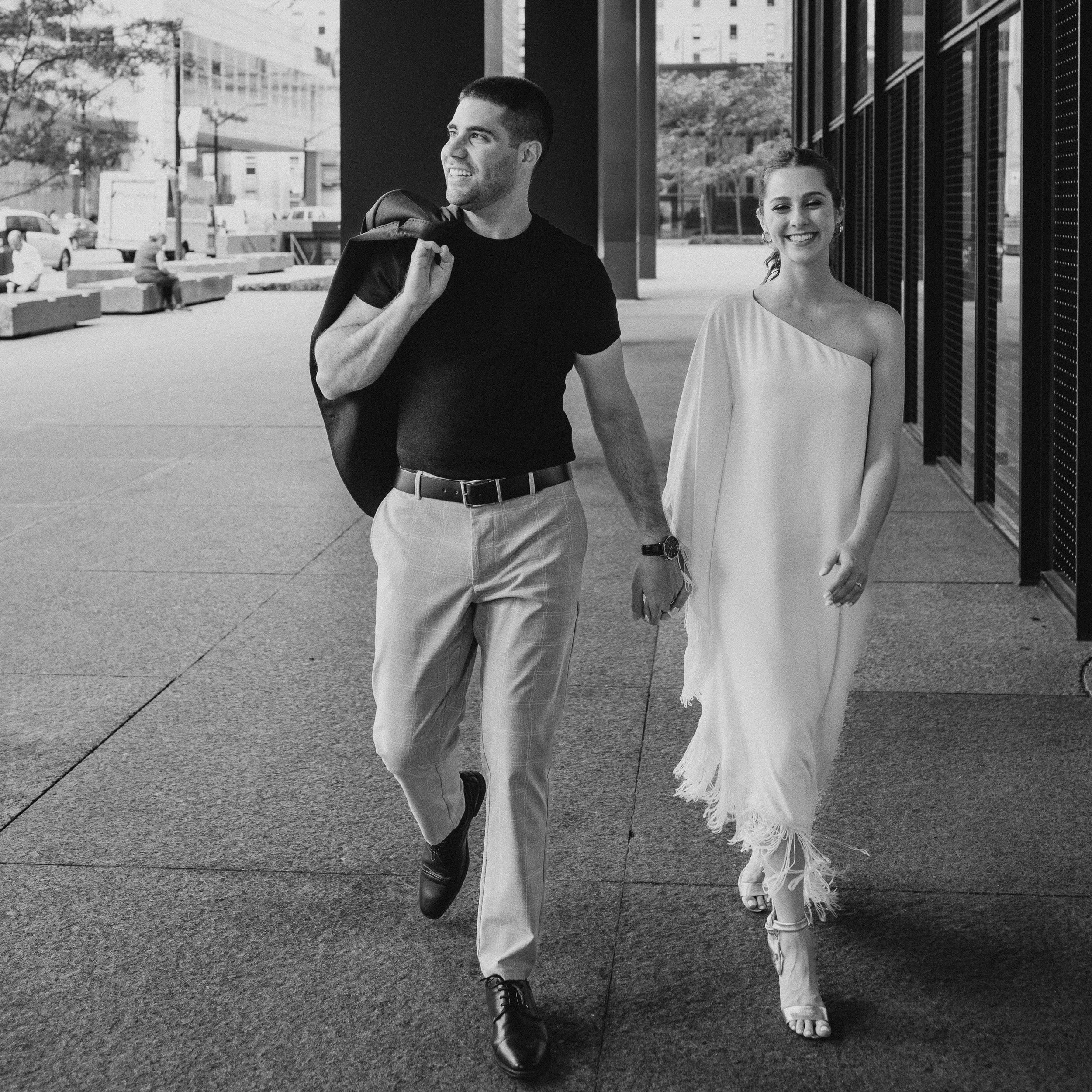 A young man and woman are walking hand in hand outside of a modern building. The man is wearing a black t-shirt and plaid trousers, carrying a blazer over his shoulder. The woman is dressed in a one-shoulder, flowing white dress with fringed hems, and is smiling. In the background, there are other pedestrians and city buildings.