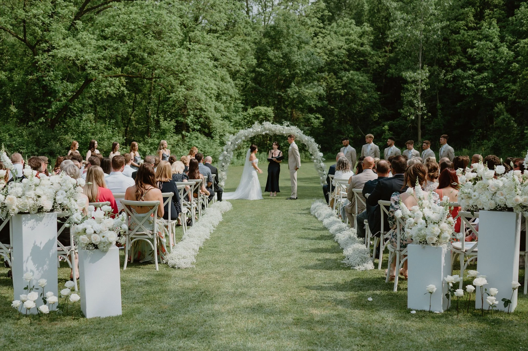 Outdoor wedding ceremony with a bride and groom standing under a floral arch, surrounded by guests seated on both sides on a lawn, with lush green trees in the background and white floral decorations along the aisle.