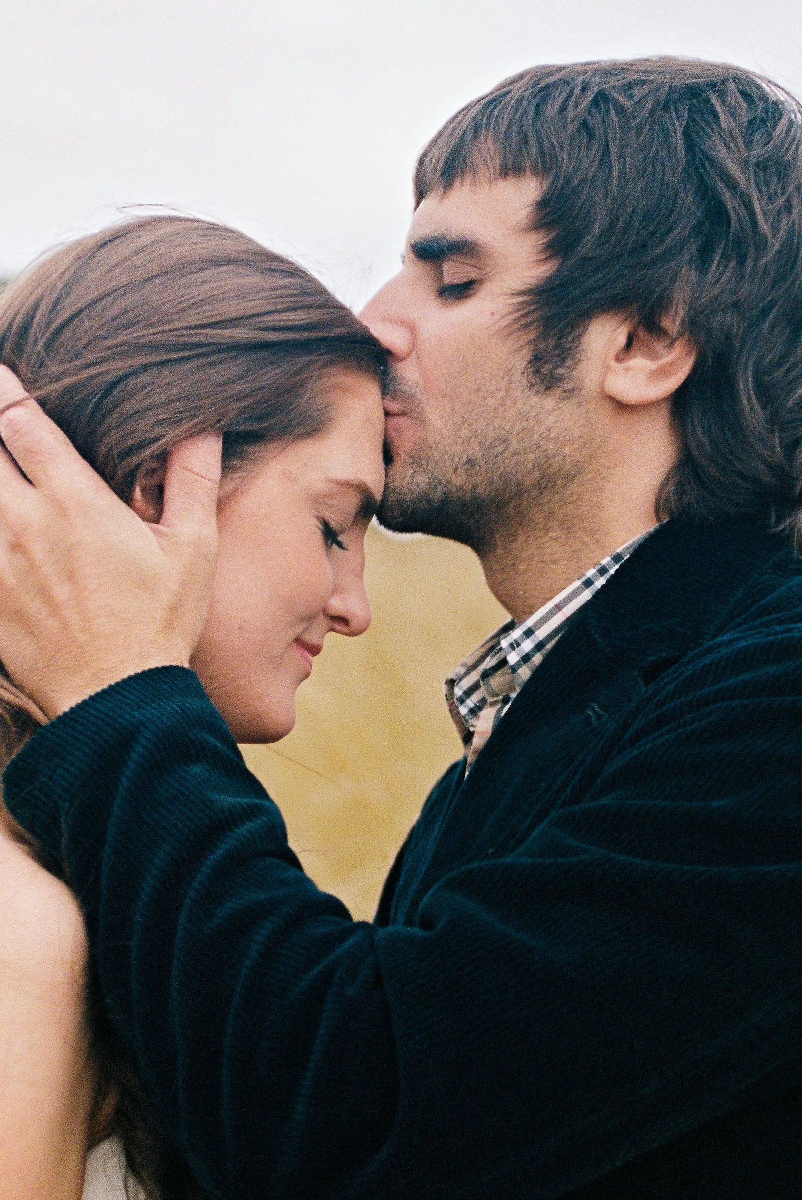 A couple embracing outdoors, with the man gently kissing the woman's forehead, both appearing peaceful.